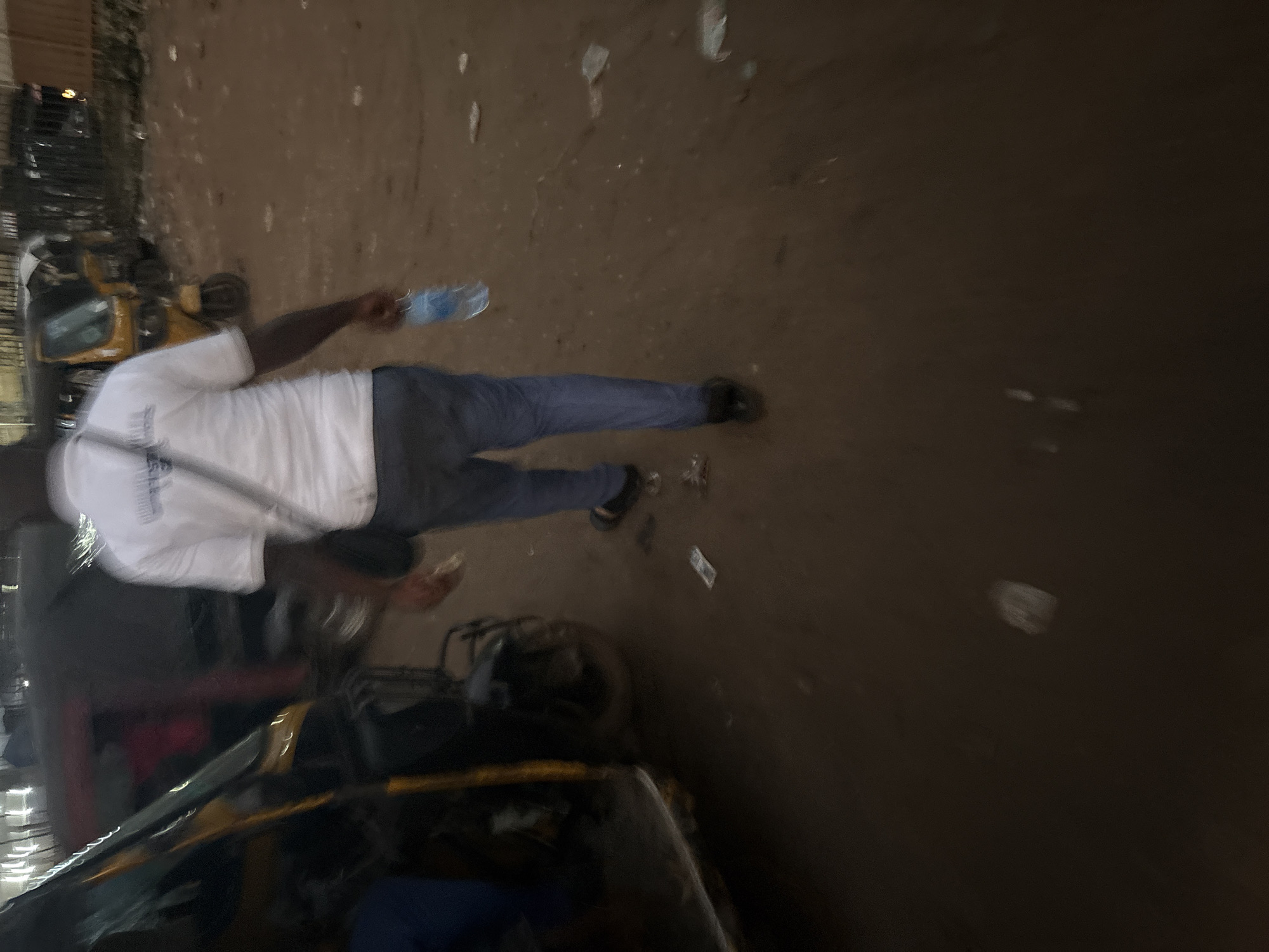 The image depicts a street scene in Onitsha, Nigeria, during what appears to be evening or dawn, given the low light conditions and artificial lighting visible in the background. In the foreground, a man wearing a white t-shirt and blue trousers walks away from the camera. He has a dark-skinned complexion and is carrying a clear plastic bottle of water in his right hand. A dark-colored bag is slung across his left shoulder.
To the left of the man, a yellow and black motorized tricycle, commonly known as an "okada" or "keke," is parked. Part of another tricycle, appearing red and dark grey, is visible behind the first. In the background, the outlines of buildings and more vehicles suggest a bustling urban environment. The ground is a mixture of dirt and what seems to be scattered debris, including some paper items. The overall impression is that of a typical street in a Nigerian city, likely in a commercial or transportation hub.