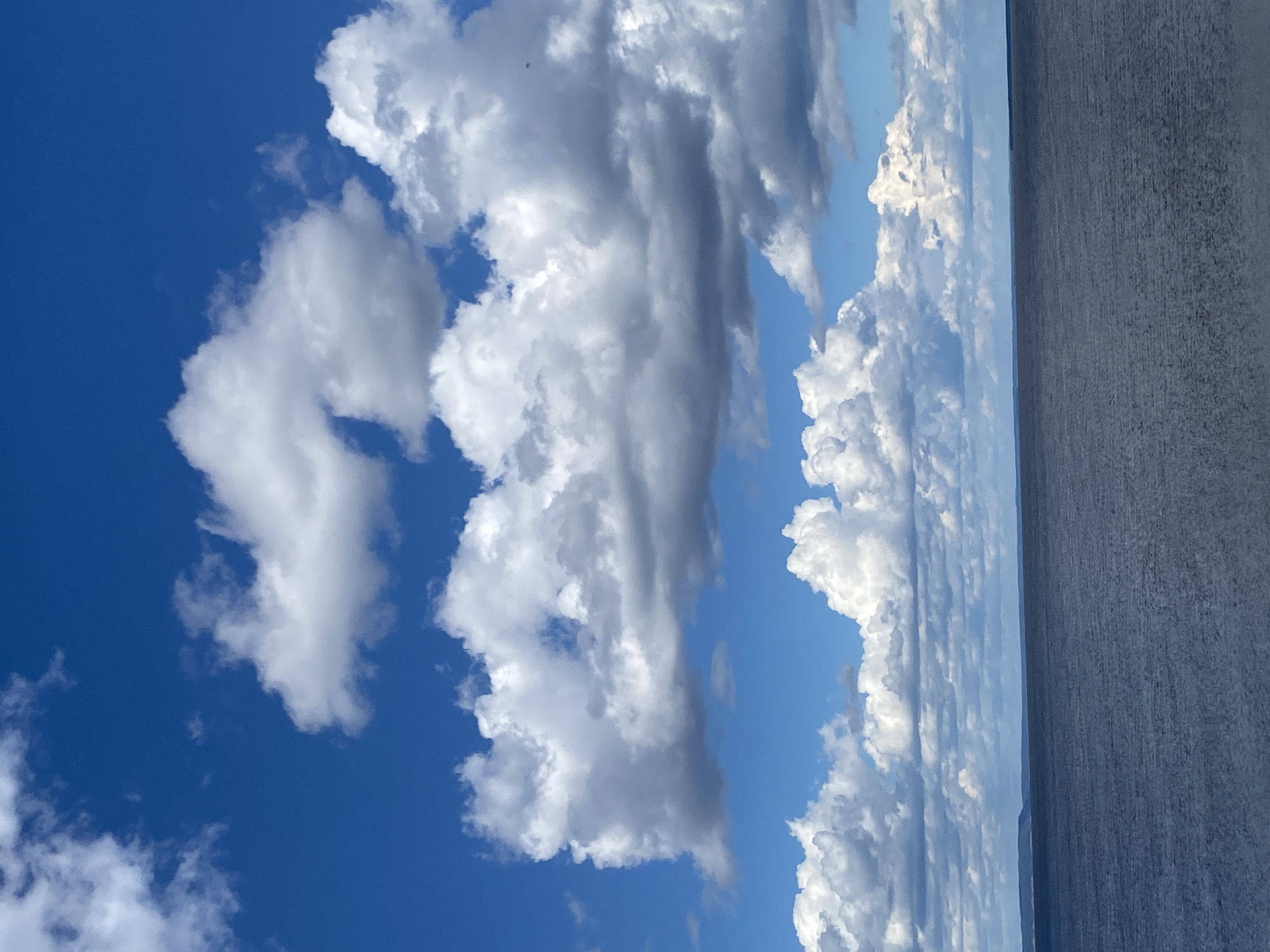 The image shows a view of the sky with white cumulus clouds against a blue backdrop. To the right of the cloudscape, there appears to be a large body of water. On the far right is a gray structure, possibly a building or a concrete wall. The image is oriented sideways. Given the specified location of Chaikovsky, Russia, the body of water could be part of the Votkinsk Reservoir. There are no visible people or interactions depicted in the photograph.