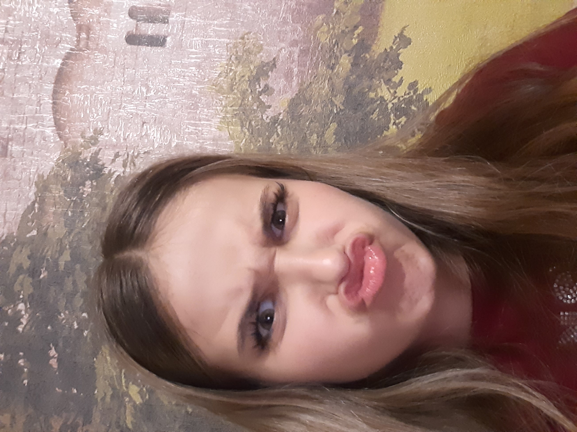 This is a close-up, rotated image of a young person making a fish-face expression. The subject has long, light brown hair and is wearing a dark red top with some sparkly embellishments visible on the shoulder. Their eyes are wide open, and their lips are pursed. The background appears to be a textured wall with a patterned wallpaper. The lighting suggests an indoor setting, possibly at any time of day. The overall emotion conveyed is playful or humorous due to the exaggerated facial expression. There is no visible text in the image.