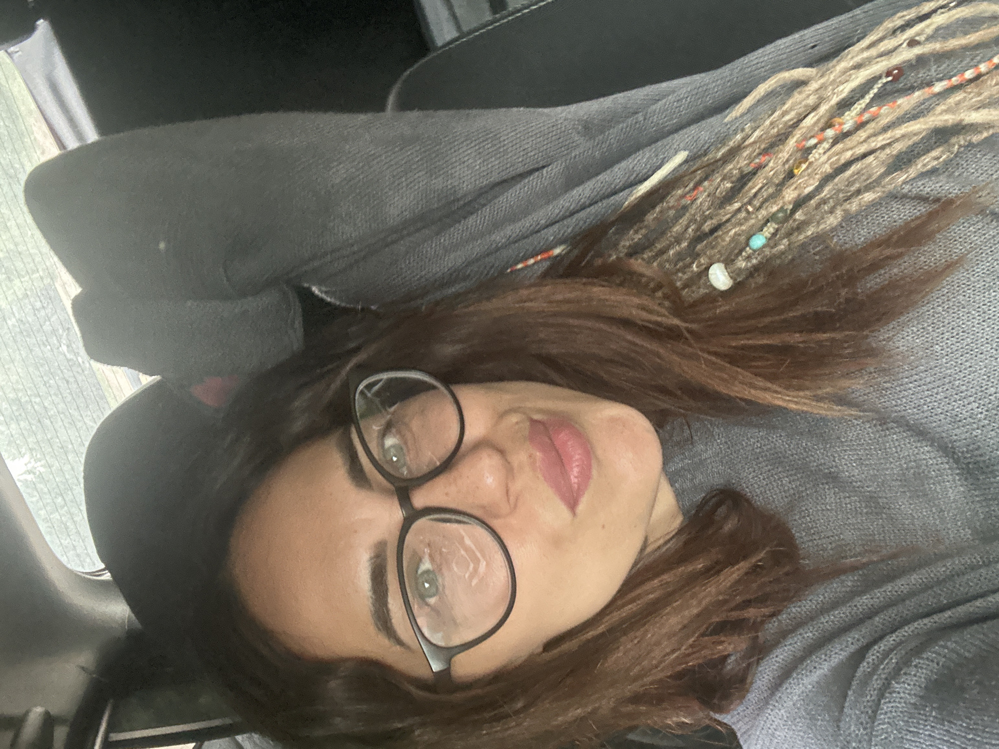 A woman lies on a car seat. She has brown hair with blonde dreadlocks decorated with beads. She wears black-framed glasses and a gray sweater. The car is likely in Chaikovsky, Russia, although this is not explicitly visible in the image.