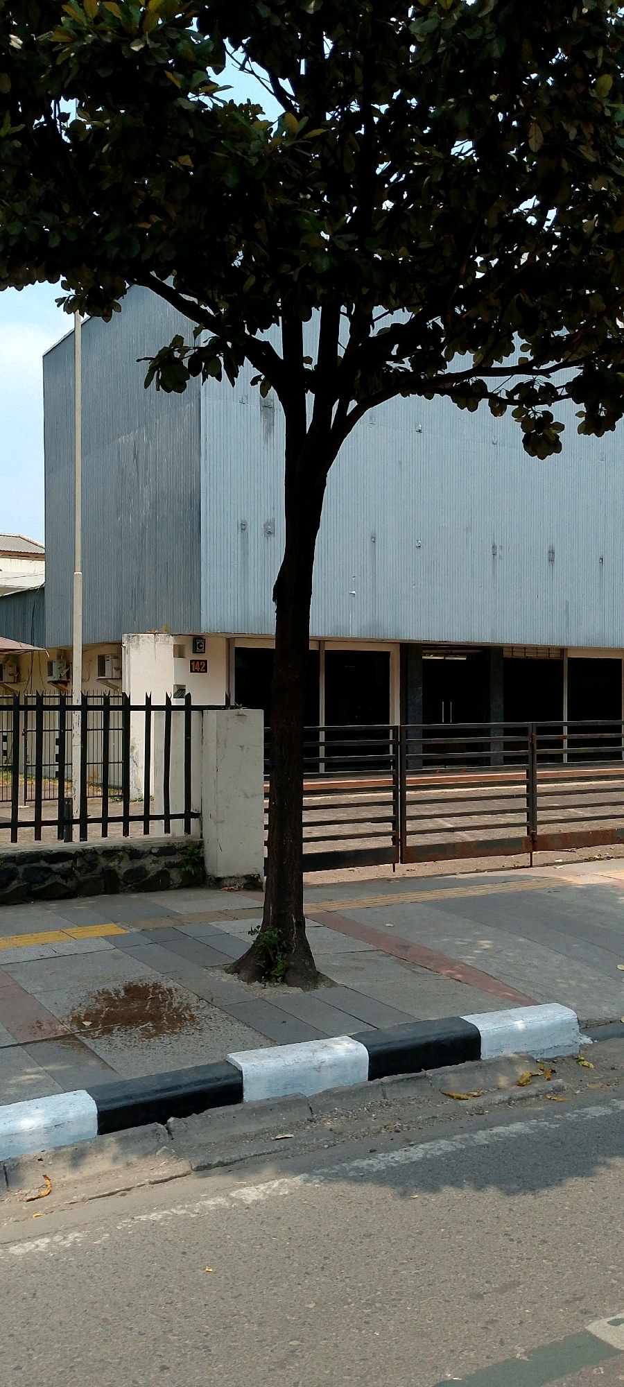 The image depicts a street scene in Bandung City, Indonesia, featuring a large, light blue corrugated metal building with a dark-tinted window facade. In the foreground, a dark tree trunk with sparse dark green foliage stands in front of the building, casting shadows. The ground is paved with concrete tiles and a striped curb painted white and black. A black metal fence with pointed finials separates the building's property from the sidewalk. A small white concrete pillar with the number "142" and a green circle logo is visible near the fence. The street itself is asphalt, with a white line marking a lane. A small puddle of water is present on the sidewalk near the base of the tree. The sky is visible in the upper background, appearing overcast.