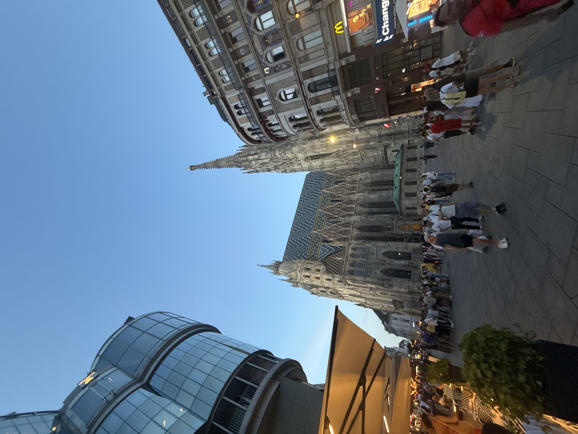 The image captures a vibrant street scene in Vienna, Austria, likely during the late afternoon or early evening given the warm, low-angled sunlight casting soft shadows. The setting is an urban plaza, bustling with activity and featuring a mix of historical and modern architecture.
Dominating the skyline is the impressive Gothic architecture of St. Stephen's Cathedral, its intricate spires and ornate facade clearly visible, topped with a distinctive, colorful tiled roof. Adjacent to the cathedral, a more contemporary building with a curved, glass facade adds a modern contrast to the historic surroundings.
A large crowd of people is the primary subject, suggesting a popular tourist spot or a gathering event. They are seen walking, standing, and milling about the plaza, many appearing to be tourists taking in the sights. Their attire suggests warm weather, with many wearing light clothing. A McDonald's sign is visible on one of the buildings, a familiar global brand in a historic European city. Text on a sign reads "Change Money".
The overall mood is lively and energetic, indicative of a busy urban environment. The clear blue sky suggests fair weather. The composition, with its dramatic upward angle, emphasizes the grandeur of the architecture.