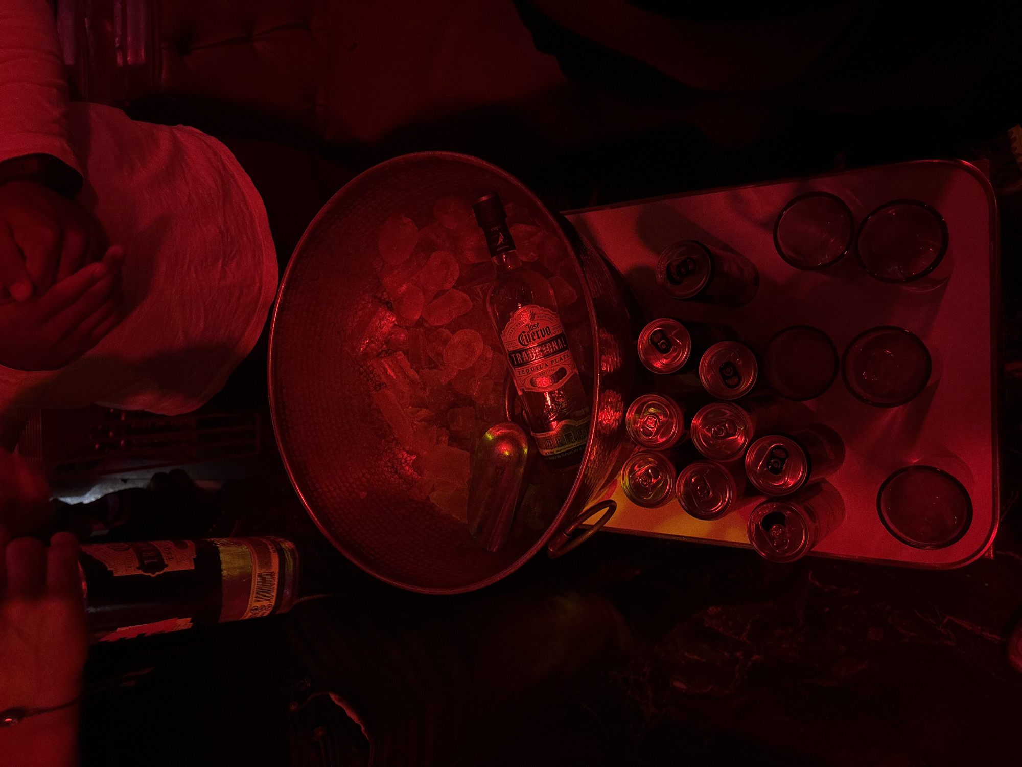 In a dimly lit environment in Puebla, Mexico, a person in a white shirt sits next to a metal ice bucket containing a bottle of Jose Cuervo Tradicional tequila and ice. Next to the bucket, a serving tray contains multiple cans and glasses, which appear to be empty. A beer bottle sits on a dark surface to the left. The scene is primarily illuminated with red light.