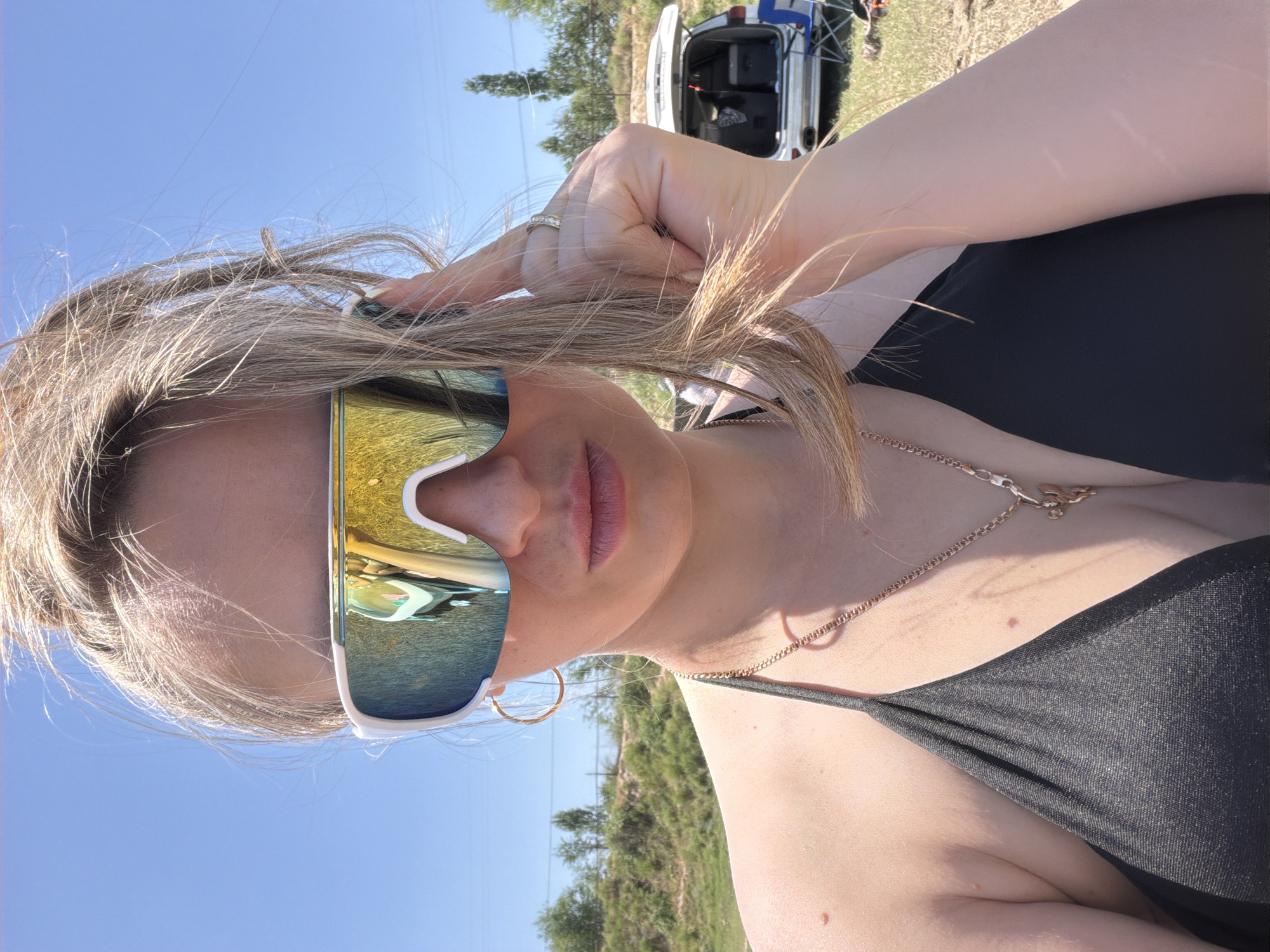 A medium shot captures a young woman in a black bikini, looking directly at the camera with a pout. She is wearing large, white, mirrored sunglasses that reflect a colorful, abstract image of the environment. Her blonde hair is pulled back into a high ponytail, with some strands falling around her face. She is touching her hair with her right hand, revealing a gold ring on her finger. A thin gold chain with a pendant is visible around her neck. The background suggests an outdoor setting with green grass, trees, and a clear blue sky. A white SUV with its trunk open, containing coolers and other items, is parked in the background, along with a blue camping chair. The scene appears to be a sunny day. The location in the city of Shymkent, Kazakhstan is not discernible from the provided image.