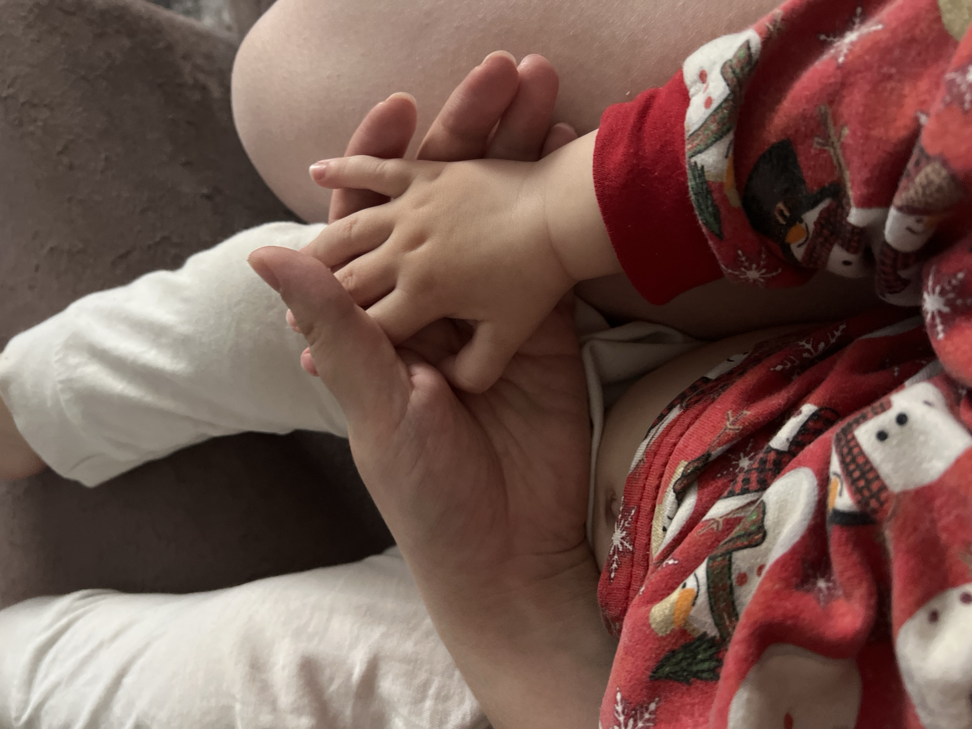 A close-up shot shows a person's hand holding a toddler's hand. The toddler is wearing red pajamas with snowman designs. The hands are positioned on a surface covered with white fabric. The environment appears to be indoors. There is no evidence to support that this image was taken in the city of Noviy, Russia.