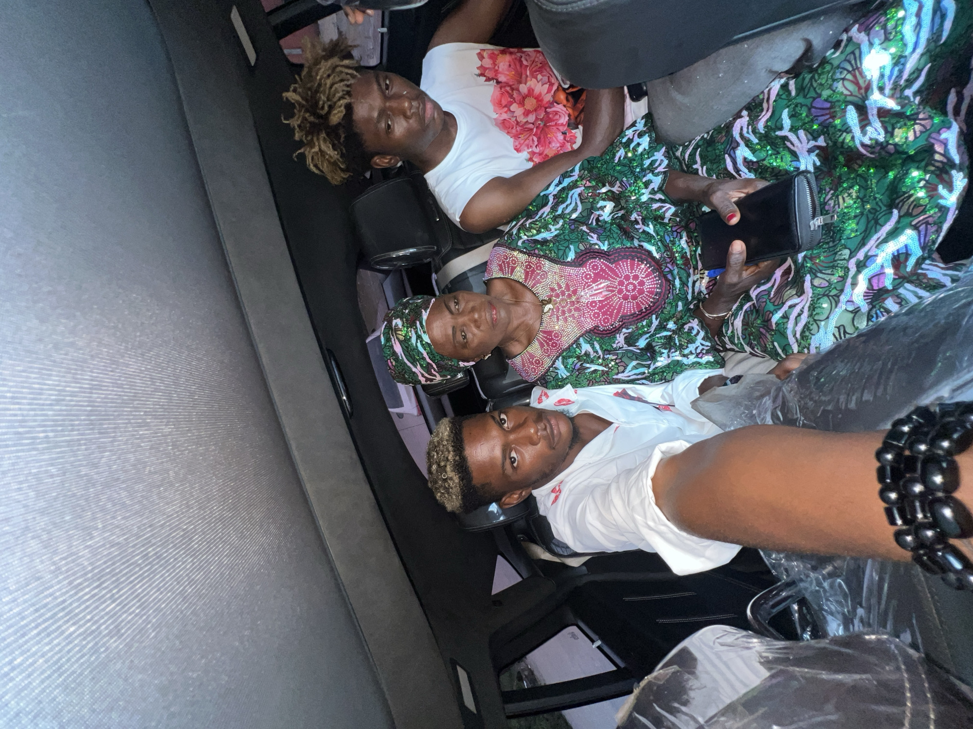 Three individuals are in a car. The first person is an older woman wearing a green, pink, and white sequined dress and a matching head covering. She is holding a black purse or wallet. The second person is a young man in a white t-shirt with a floral design. The third person is a young man wearing a white shirt and a black beaded bracelet, who appears to be taking the picture. Based on the context and the individuals pictured, it is likely that this image was taken in Igboefon, Nigeria. The car's interior appears to be gray.