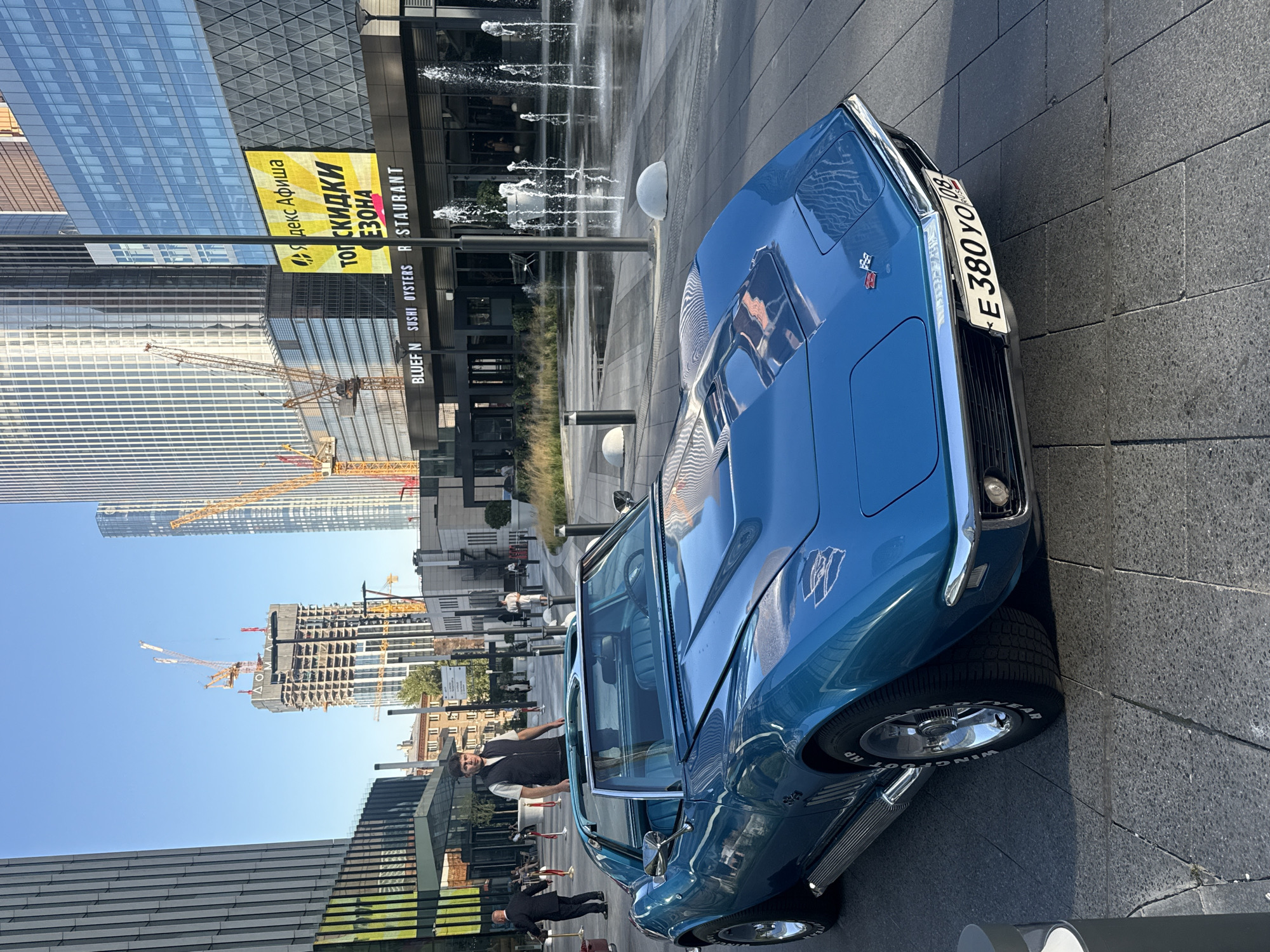 A bright blue 1960s Chevrolet Corvette Stingray is parked on a stone-tiled plaza in front of a modern building with large glass windows and a rooftop fountain. The car's convertible top is down, revealing blue interior seats. A man in a dark vest over a white shirt stands near the car, looking towards the camera. In the background, several tall, contemporary buildings are under construction, with yellow cranes visible against a clear blue sky. A large yellow sign with Russian text, which appears to advertise discounts and events, is mounted on the building facade above a restaurant with the name "BLUEFIN SUSHI OYSTERS RESTAURANT" clearly visible. The overall scene suggests an urban environment, likely a business or entertainment district in Khimki, Russia, possibly during daylight hours due to the bright lighting and shadows.