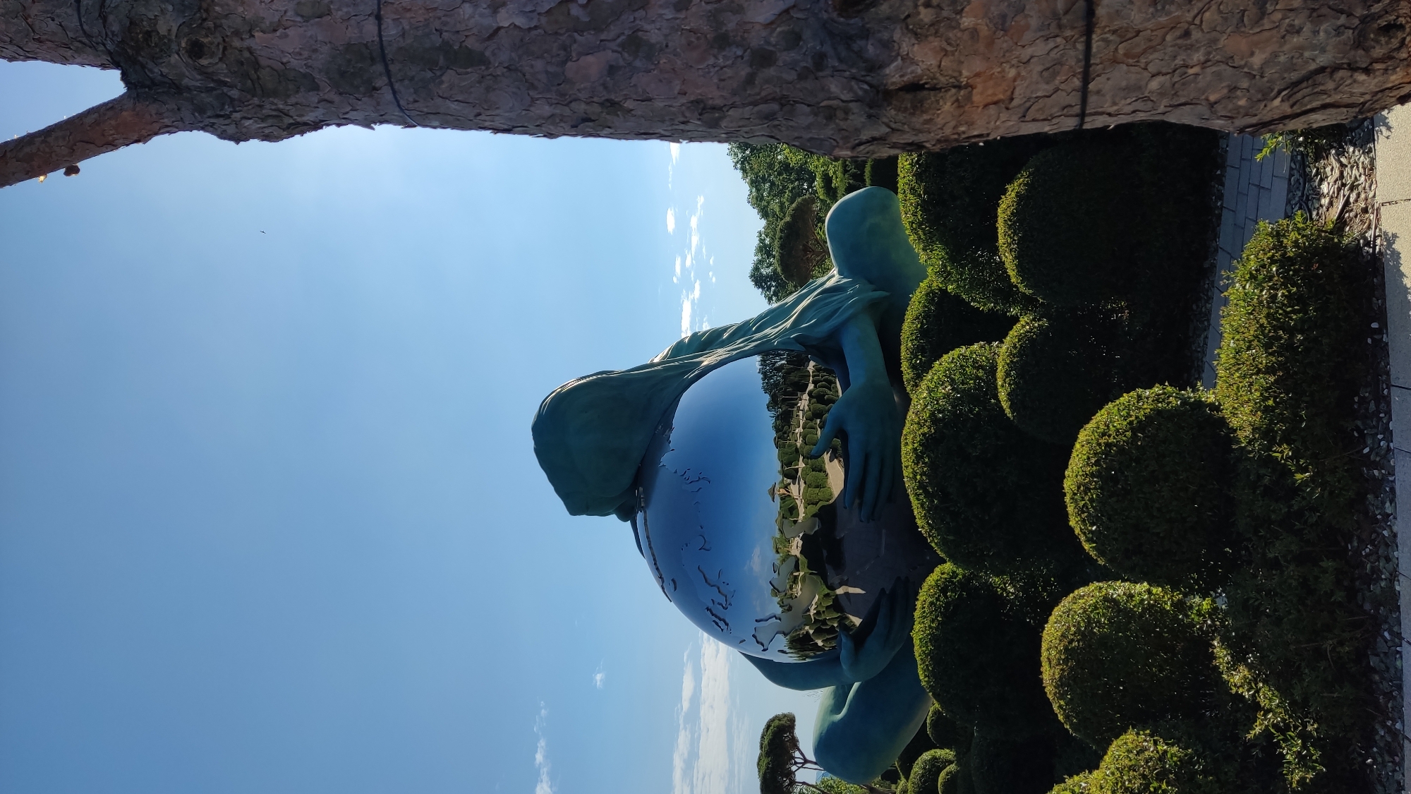 A large, reflective sculpture of the Earth is cradled by a stylized, teal-colored figure with a hooded head, suggesting a maternal or protective pose. The sculpture is situated outdoors in a garden setting, surrounded by manicured, spherical bushes and other topiary forms. In the background, a line of trees can be seen, and beyond them, a bright blue sky with scattered white clouds. The reflection on the Earth sculpture reveals the surrounding garden, including what appears to be a gazebo-like structure and more greenery. To the right of the frame, a portion of a large tree trunk with textured bark is visible. The scene is brightly lit, indicating daylight. This artwork is located in the "Galitsky Park" in Krasnodar, Russia.