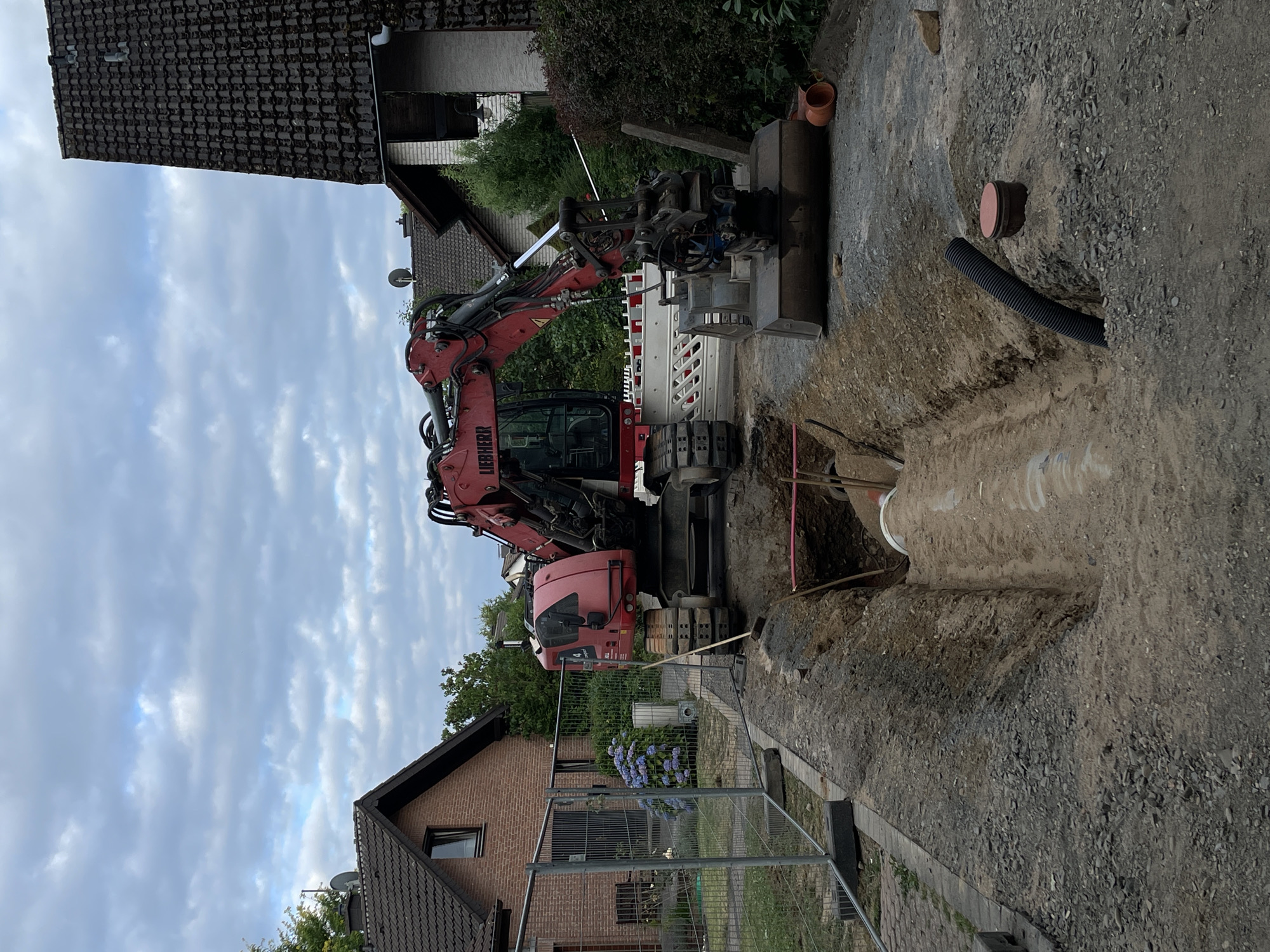 This image depicts a construction scene in Hennef, Germany. A large red Liebherr excavator is positioned next to a freshly dug trench where a large pipe is being installed. The trench is filled with soil and gravel. To the left of the trench, a temporary metal fence encloses a paved area and a brick house with blue hydrangeas in front. In the background, more residential buildings and trees are visible under a cloudy sky. There are no people visible in the image. The scene appears to be part of a utility or infrastructure project.