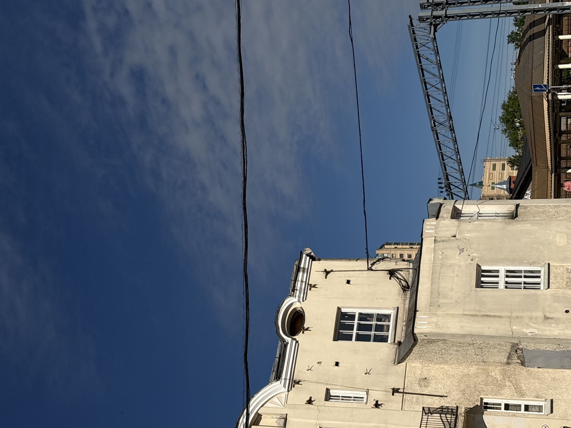 The image, taken with an upward perspective, captures an urban sky view over Saint Petersburg, Russia, on a bright day. The clear blue sky, partially adorned with sparse, wispy white clouds, dominates the upper and left portions of the frame.
In the foreground, a prominent light-colored building, possibly beige or off-white, fills the right side of the image. Its facade displays a blend of architectural styles, including a distinctive large, circular, recessed window (oculus) positioned within an ornate, curved architectural element. Below and to the right of this, several rectangular windows with white frames and horizontal mullions are visible. The building shows signs of age and wear.
Stretching across the sky horizontally are multiple thin, dark power lines or cables. In the upper-left, a large, grey metal truss structure, characteristic of an overhead railway or metro line, extends across the frame. Beneath this structure, in the midground, is another building topped with a brown awning or roof, and some green trees or bushes are visible behind it. A small, blue and white road sign is also present near this brown structure, though its text is not legible. Further in the background, another light-colored building with multiple windows can be seen.
The scene suggests a sunny daytime, likely morning or afternoon, given the bright, direct light. No specific activity or event is taking place, and there are no people visible. The image conveys a sense of an established urban environment with a mix of historical architecture and modern infrastructure.