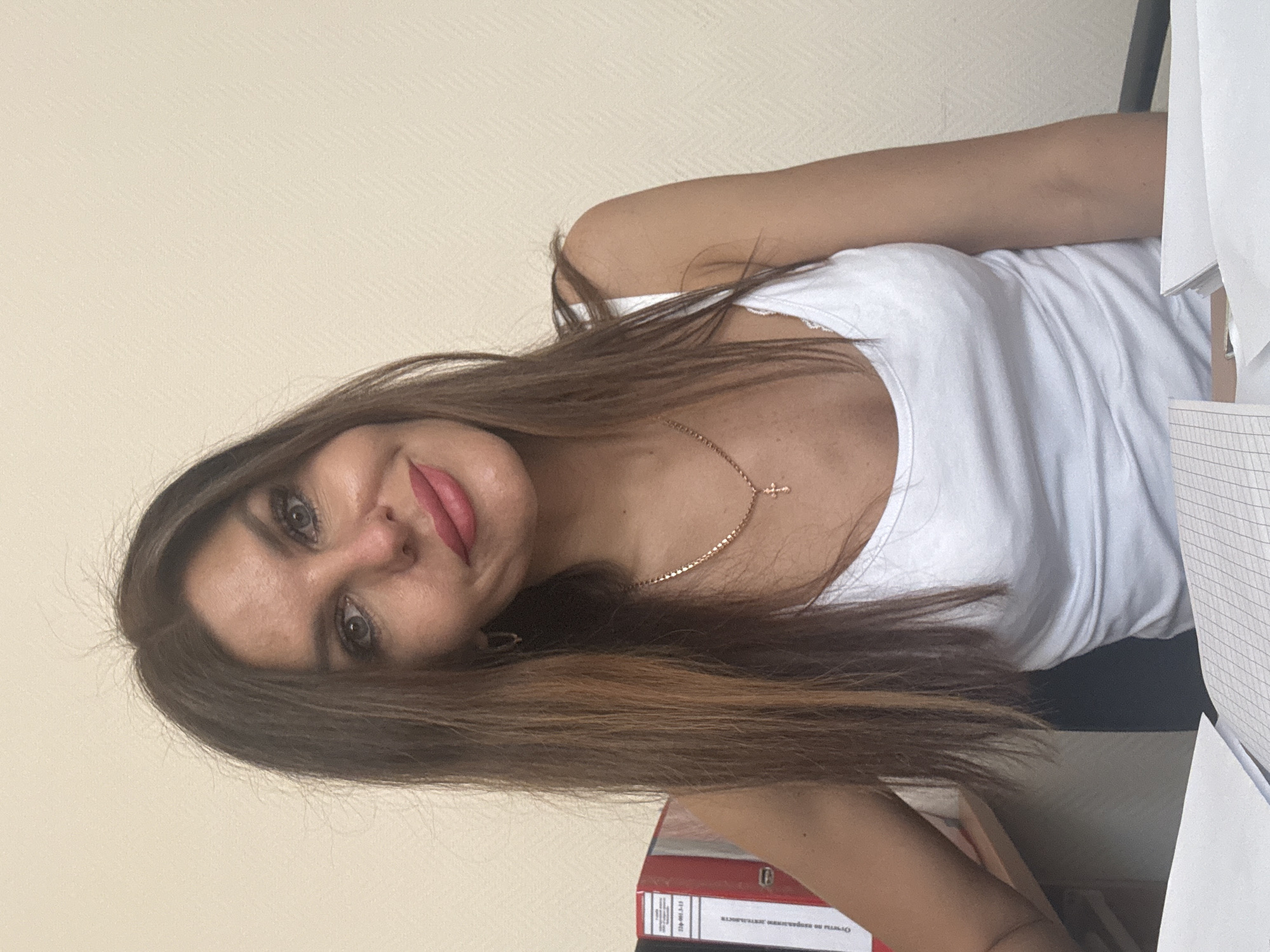 A young woman with long brown hair is posing for a selfie indoors. She is wearing a white tank top and a gold necklace with a cross pendant. She is smiling with red lipstick. A notebook and papers are visible in the frame, along with a folder. While the location is stated as Chaikovsky, Russia, there is nothing visible in the image that confirms that location.