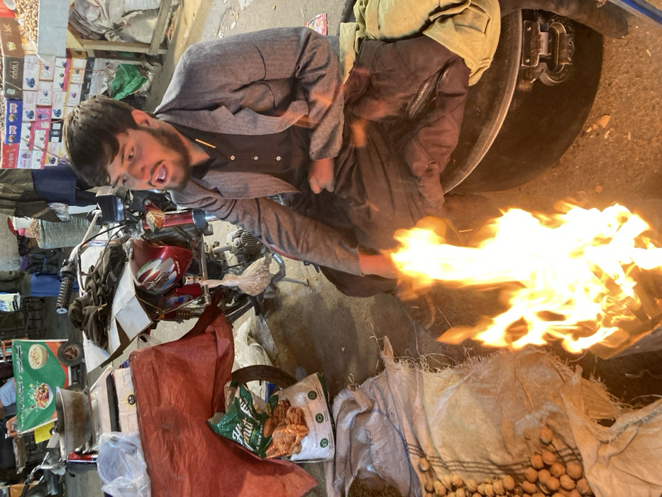 A man in a gray jacket and dark pants sits beside a motorcycle, his hand near a large flame. He appears to be yelling or speaking. Next to him is a large sack filled with nuts or seeds. The background shows a cluttered scene, possibly a market, with boxes of products, fabrics, and various objects scattered around. The image has been rotated slightly, but the overall impression is one of activity and chaos.