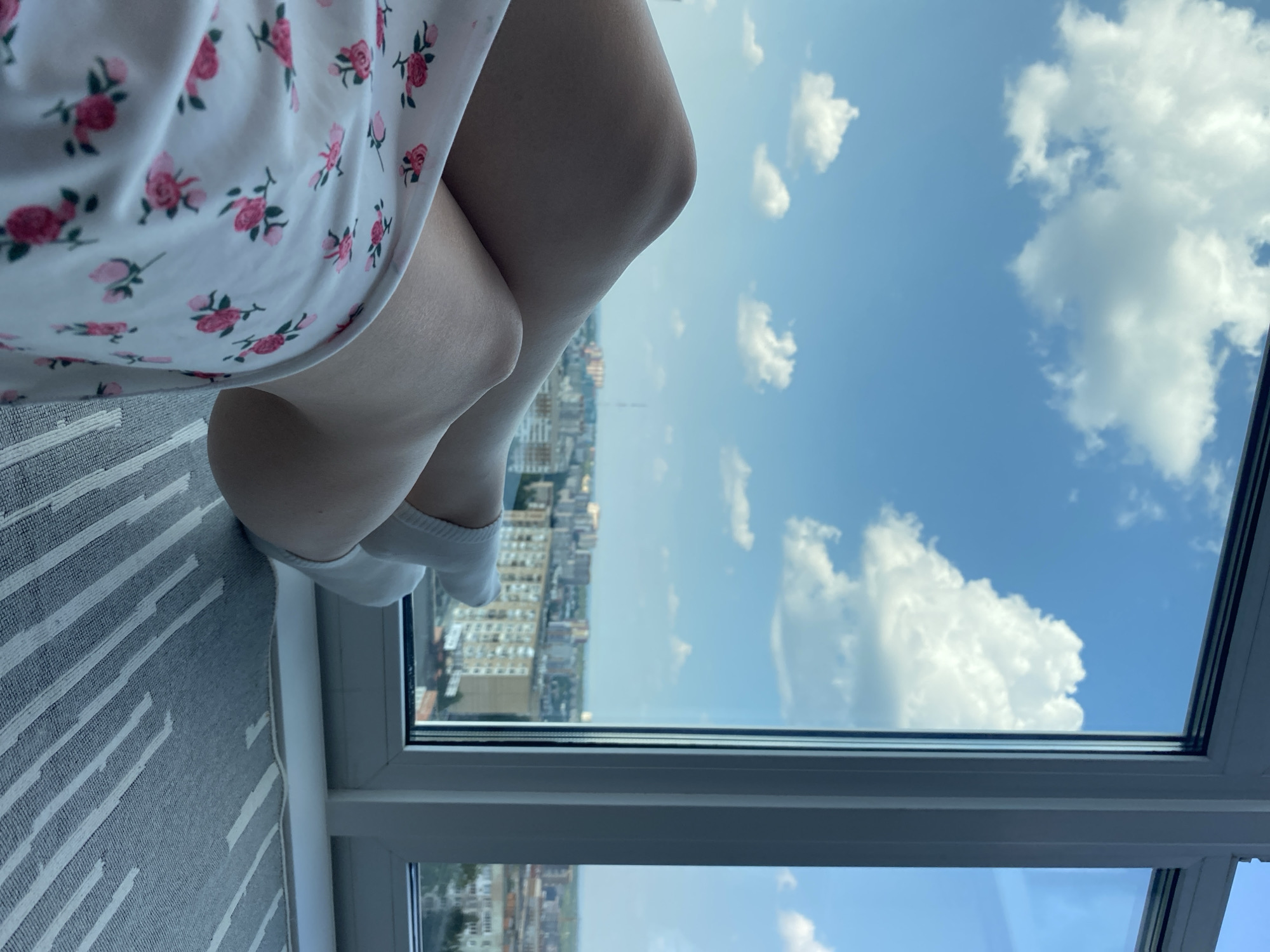 A close-up shot focuses on a person's legs, clad in white socks and a floral-patterned dress, resting in front of a large window. Outside the window, a cityscape unfolds, featuring buildings and a hazy horizon under a bright blue sky dotted with white clouds. The scene is likely from an apartment in Izhevsk, Russia, given the architectural style of the buildings. The foreground shows a portion of a rug with a striped pattern. The lighting suggests it is daytime, possibly late morning or early afternoon.