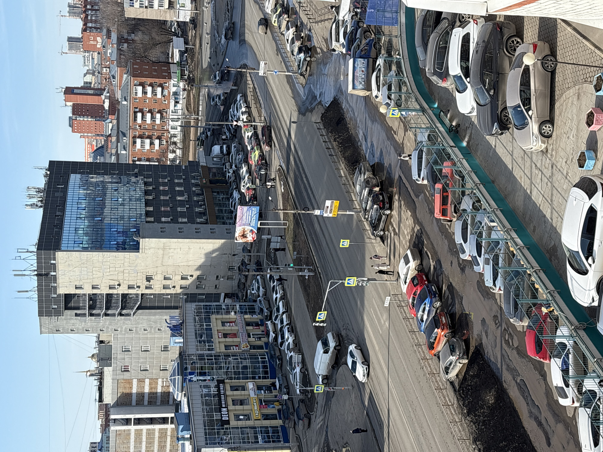 The image is an overhead shot of a city street in Izhevsk, Russia. The buildings are a mix of high-rise residential and commercial structures. The street is divided with two lanes of traffic going each way, and there are crosswalks at regular intervals. Several people are walking on the sidewalks and crossing the street. Cars line both sides of the street in designated parking spots. Some of the parking spaces have barriers around them. The sky is clear and blue, and the overall impression is one of a busy city street.