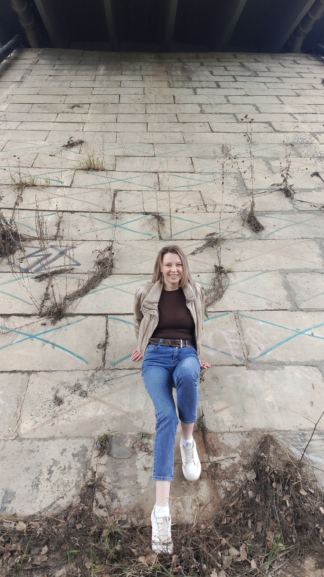 A cheerful woman is pictured sitting on a concrete retaining wall in Chaikovsky, Russia, smiling directly at the camera. She has shoulder-length light brown hair and is dressed in a light brown jacket, a dark brown top, blue jeans, a brown belt, and white sneakers. Her legs are bent, with her feet resting on the dry ground and sparse foliage at the base of the wall.
The background is dominated by a tall, textured wall composed of large, weathered grey concrete blocks. The wall shows signs of age, with cracks and some dry weeds or grass growing from them. Blue linear markings, possibly chalk or paint, crisscross parts of the wall. To the woman's left, graffiti including a distinct "W" shape is visible. Above the concrete wall, dark structural elements and pipes of what appears to be a bridge or overpass can be seen, indicating the setting is beneath an elevated structure. The overall impression is daytime, with diffused lighting, suggesting an overcast day or a location shadowed by the overhead structure. The sparse, dry vegetation points towards a non-growing season, such as late autumn, winter, or early spring.