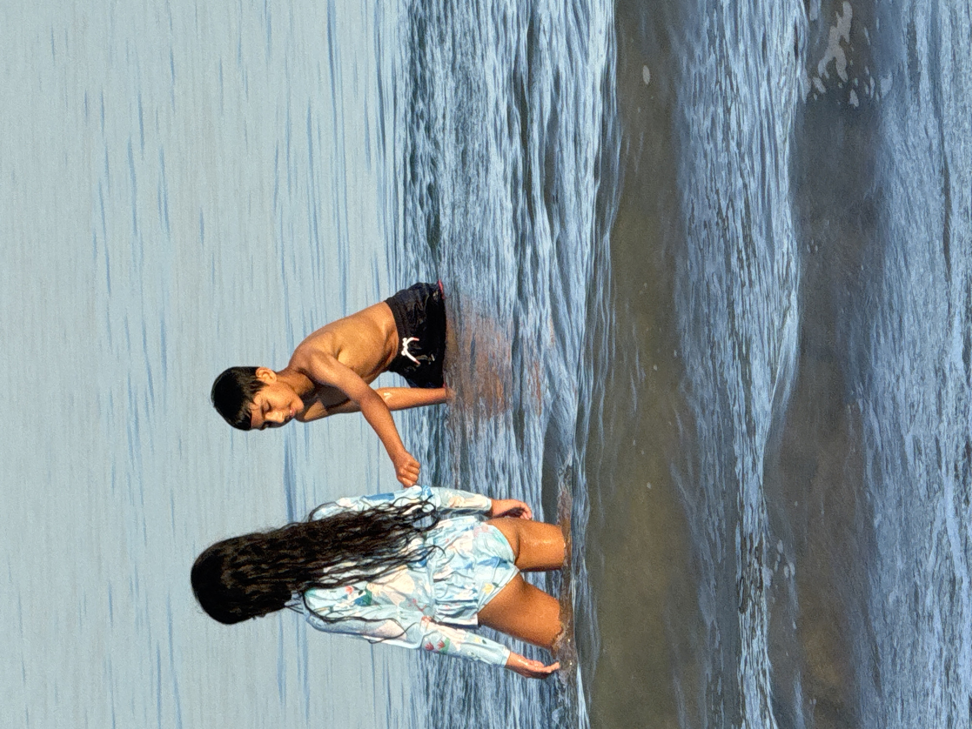 Two children stand in shallow water at a beach in Jimbaran, Indonesia. A boy stands above a girl, their bodies partially submerged. The scene is shot with a rotated orientation, making the water appear vertical. The water shows ripples and depth variations. The sky is overcast and light blue, providing an even light source.