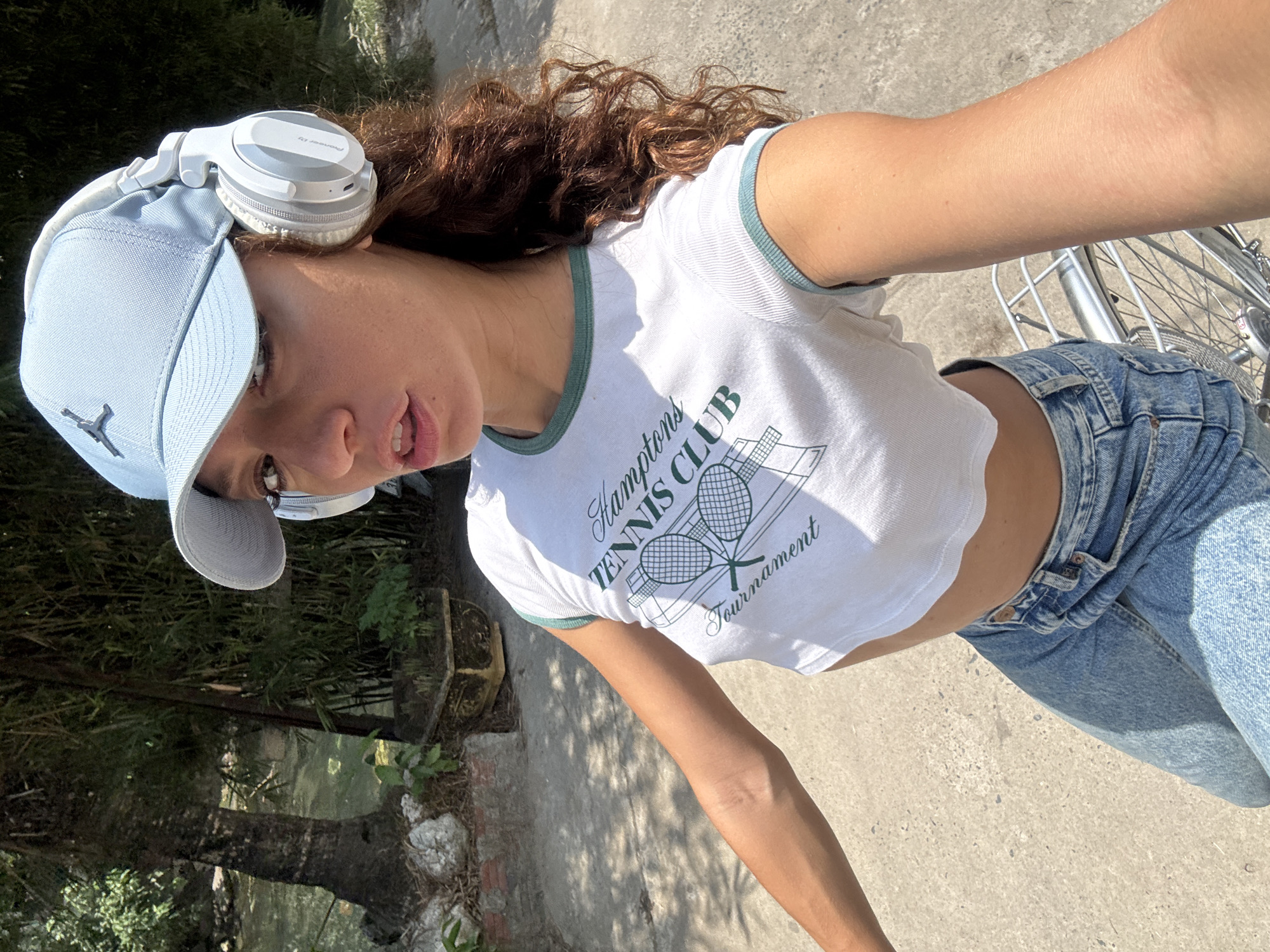 A young woman stands in what appears to be a city park or residential area with lush vegetation, possibly in Đà Nẵng, Vietnam, though this cannot be verified without further information. She is wearing a light blue baseball cap, white headphones, a white cropped T-shirt with a green trim and a "Hamptons Tennis Club Tournament" logo, and light-wash jeans. She is holding a bicycle. The surroundings include a concrete pathway, trees, and possibly other plants.