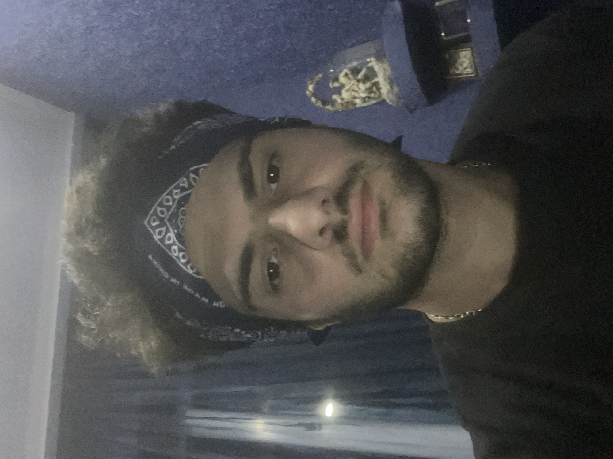 A man with curly, dark hair and a bandana is taking a selfie in a room. He is wearing a black t-shirt and a silver chain necklace. The background is a blue wall and blue curtains. To the right of the man, there is a small decorative figurine on a shelf. The lighting is dim, suggesting it may be nighttime. The image does not provide any information about the location being in Tbilisi, Georgia.