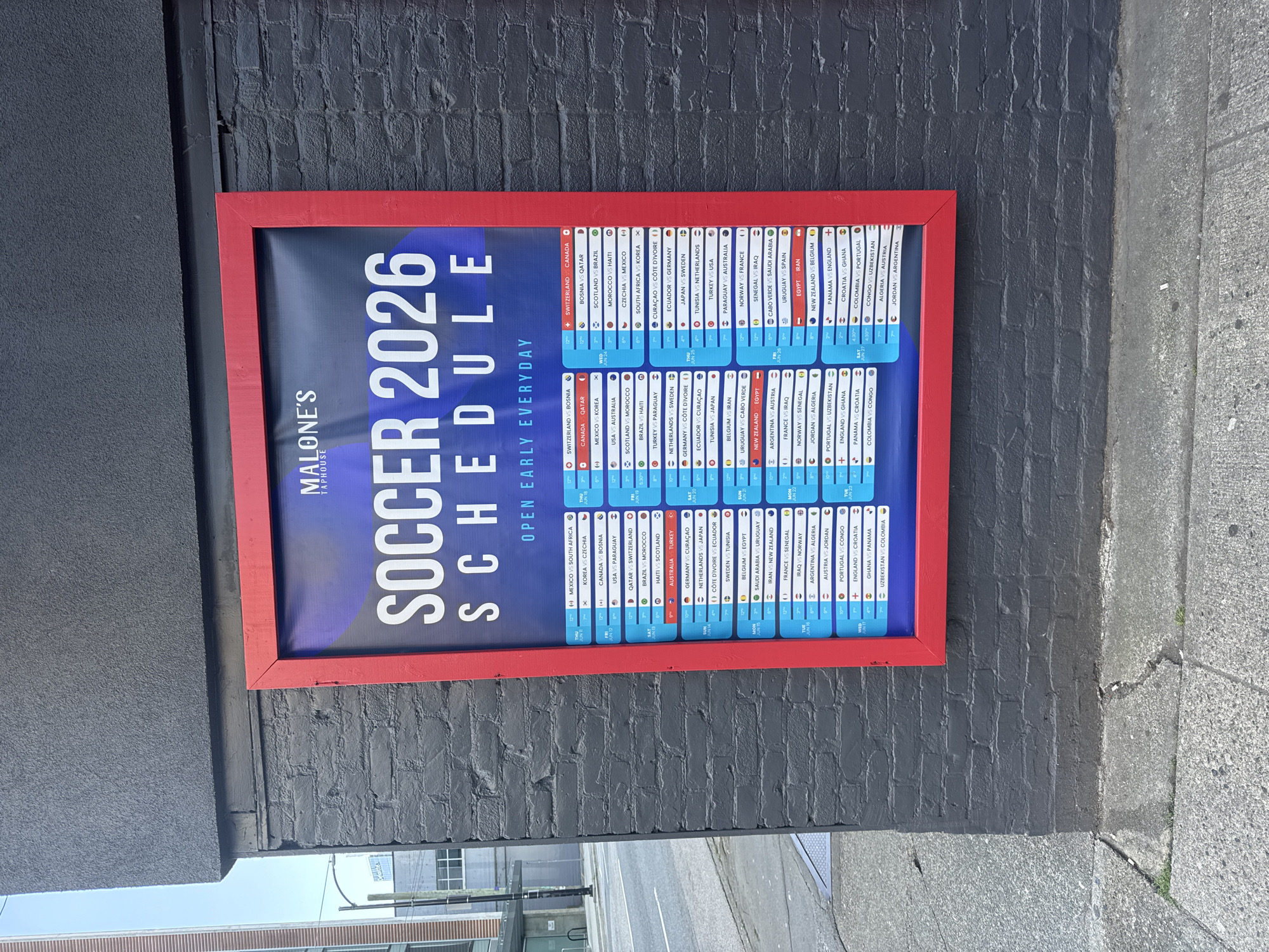 An outdoor poster advertising the "SOCCER 2026 SCHEDULE" is displayed on a dark grey brick wall. The poster has a red frame and features a dark blue background with purple accents. The title "SOCCER 2026 SCHEDULE" is in large white letters. Below the title, it states "OPEN EARLY EVERYDAY." The schedule itself lists various soccer matches with dates, times, and participating countries. To the left of the poster, a light-colored building with vertical ribbed siding and windows is partially visible. The ground in front of the wall is paved with concrete. The scene appears to be in an urban environment.