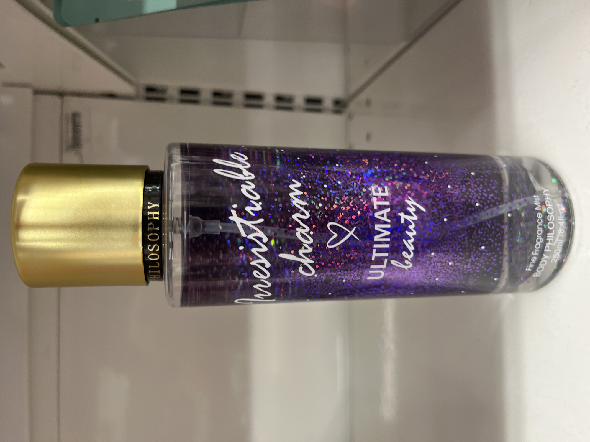 A clear glass bottle of body spray with a gold cap sits on a white shelf. The body spray is purple and filled with glitter. The label on the bottle reads "Philosophy" at the top, followed by "Irresistible charm" and "Ultimate beauty" in white lettering. The bottom of the label reads "Fine Fragrance Mist" and "BODY PHILOSOPHY" and "250ml 78.4fl.oz". The background is a blurry white shelf.