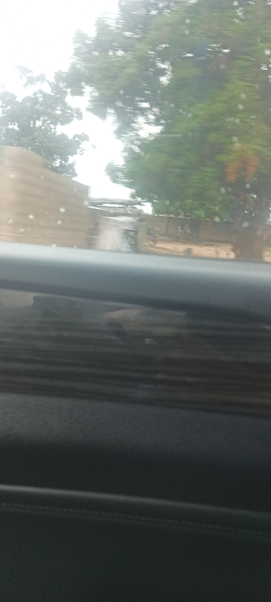 The image shows a blurred view through a vehicle window. In the distance, a dirt road leads between buildings and trees. The location is Gwadanye, Nigeria, based on the prompt. No people are visible. The foreground is the interior of the vehicle, consisting of a dashboard with horizontal lines and stitching. The weather appears overcast or rainy.