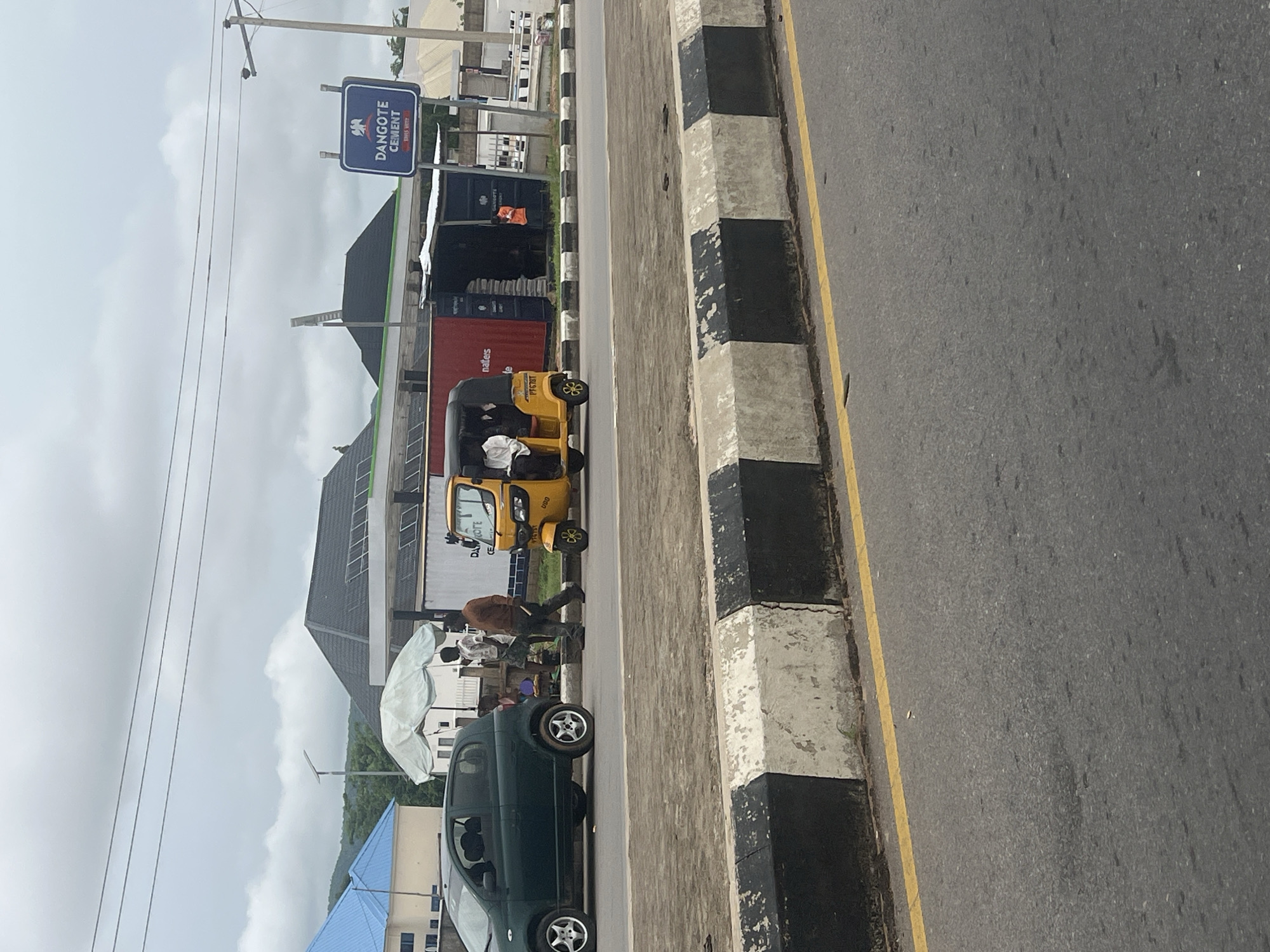 In Jalingo, Nigeria, on an overcast day, a yellow auto-rickshaw, or "Keke Napep," navigates a paved road. A dark green hatchback car is parked on the side of the road. Two men are standing near a white umbrella structure, with one man walking away from the camera. Behind them, a red shipping container is visible, with a structure built in front of it. A blue sign with "DANGOTE CEMENT" written on it is mounted on a pole to the right of the container. The road is bordered by a concrete barrier with black and white stripes. In the background, buildings with modern, dark-tiled roofs and solar panels are present, along with trees and hills. The sky is cloudy.
