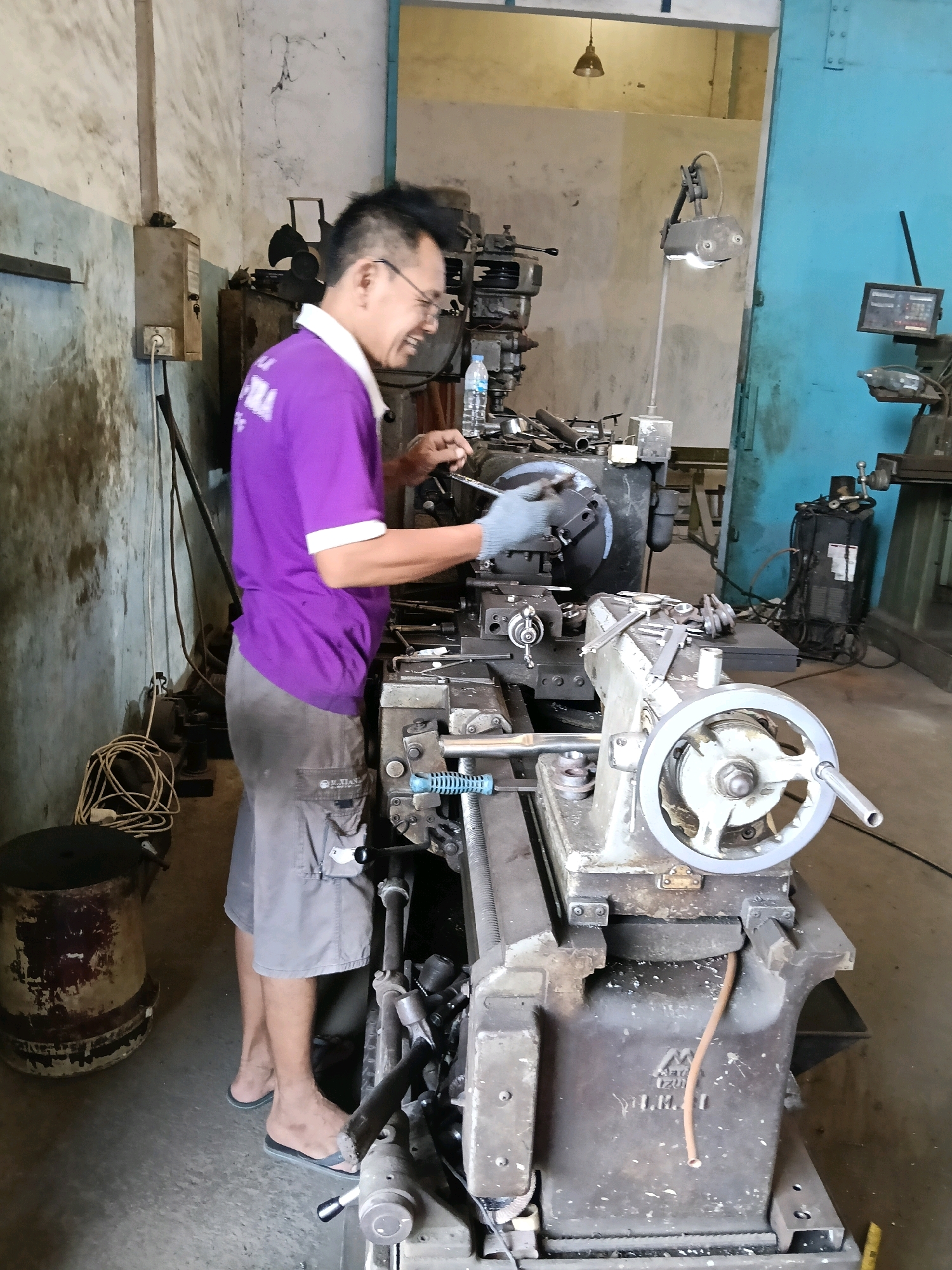 A man in a purple shirt and grey shorts operates a metal lathe in what appears to be a workshop in Belimbing, Indonesia. He wears blue gloves and glasses and is looking at the machine. Tools and metal shavings are visible on and around the lathe. The workshop is dimly lit with concrete walls and floor, and several other industrial machines are present. A water bottle is visible on the lathe.