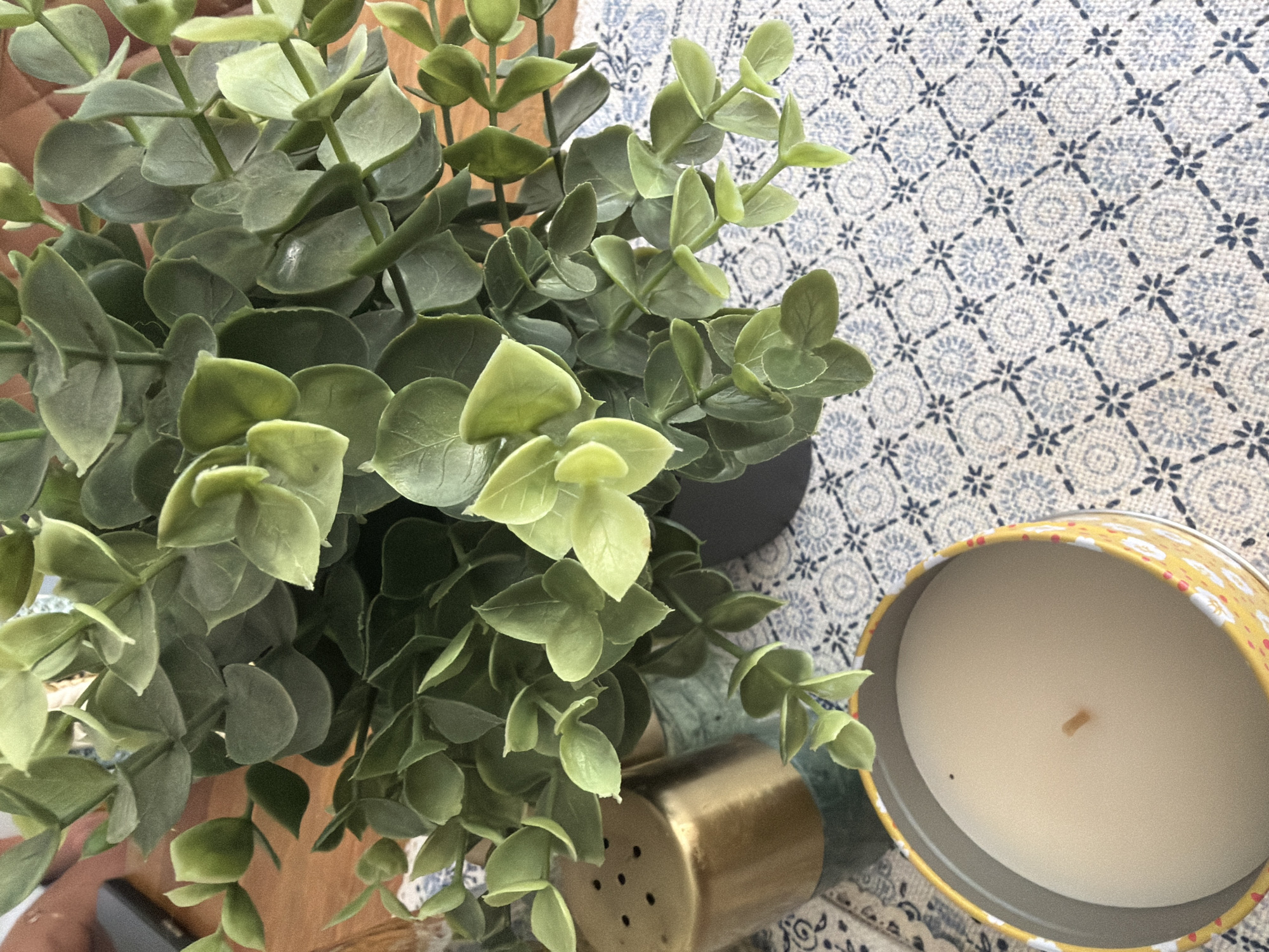 The image presents a still life arrangement on a surface covered with a blue and white patterned textile. A lush, green leafy plant dominates the left side of the frame, positioned in a dark-colored pot. Adjacent to the plant, a metallic gold object with small perforations is visible. To the right, a round candle in a yellow decorative container with a floral pattern sits on the patterned surface. The scene appears to be indoors. There is no indication of location or Guildford.