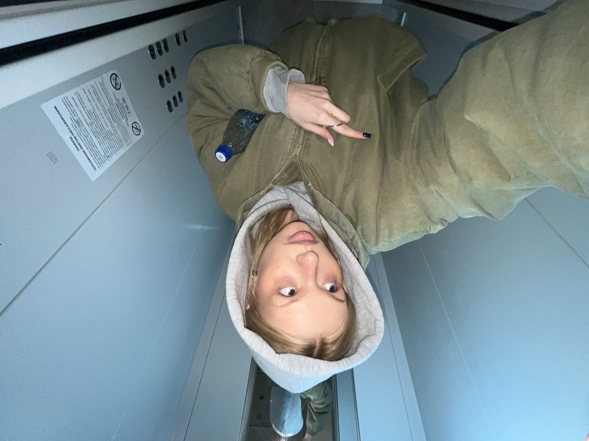 A young woman is standing inside an elevator, taking a selfie. She wears a light gray hooded sweatshirt and a light brown jacket. In the upper-left corner of the elevator, there is a sign with text written in Russian. The location is identified as Chaikovsky, Russia, based on the language of the sign. The woman is holding a plastic water bottle and displaying her middle finger. Her reflection can be seen on the elevator door.