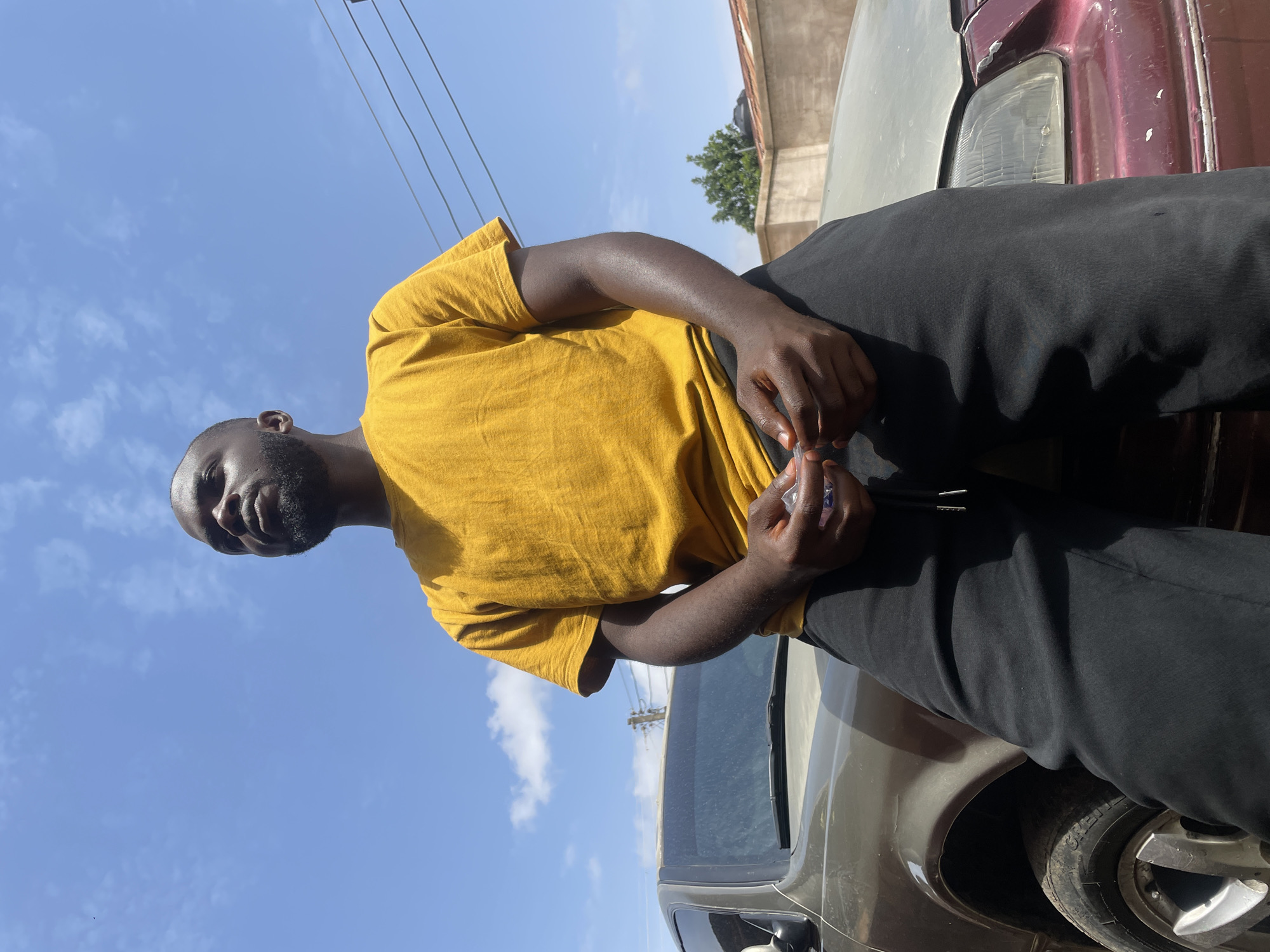 A dark-skinned man in a yellow t-shirt and black pants sits on the hood of a grey car. He holds a small, clear plastic bottle in his hand. The sky above is blue with scattered clouds. Utility lines run overhead. Parts of other cars are visible to the right and below the main subject. The background suggests a street or urban environment. The location is not explicitly discernible as Jos, Nigeria based solely on the image.
