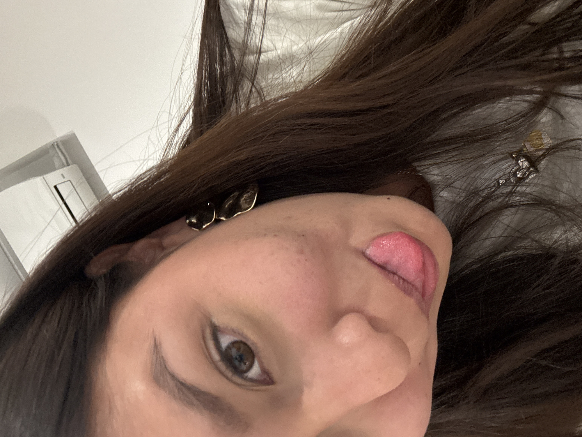 This image is a close-up, angled portrait of a single female individual located indoors in San Andrés Cholula, Mexico. The subject's head is tilted to the viewer's right, with her left brown eye directly visible, and her pink tongue is extended from her mouth. Her dark brown hair, long and straight, frames her face and covers her right shoulder. She wears a gold-toned, irregularly shaped earring on her right earlobe and a necklace with two pendants: one silver-colored in a heart shape and another gold-colored and rectangular. Her attire includes a light-colored garment, possibly a t-shirt or sweater. The background consists of a plain white wall, with a white wall-mounted air conditioning unit visible in the upper left portion of the frame.