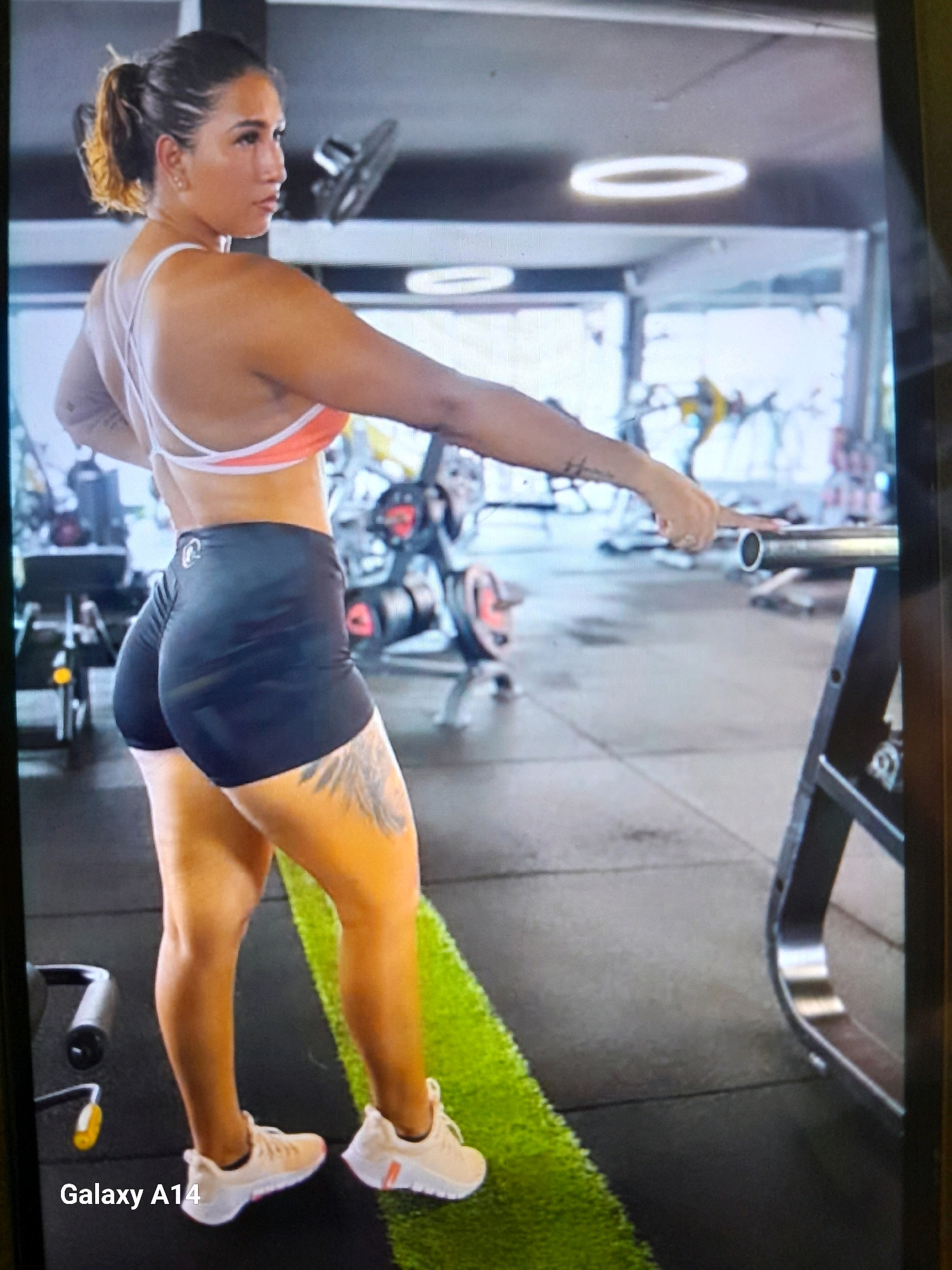 In the city of Noord, Suriname, a woman is captured mid-pose in a gym setting. She is positioned facing away from the viewer, with her left side slightly angled towards the camera. Her hair is pulled back into a ponytail. She is wearing a coral-colored sports bra with white straps and black athletic shorts. Visible tattoos adorn her left thigh. Her arms are extended, and her right hand is touching a barbell. She is wearing light-colored athletic shoes. The gym environment features exercise equipment, including weights and machines, and a bright ring light is visible on the ceiling. The floor is a mix of gray rubber mats and a strip of artificial green turf. The overall scene suggests a focus on fitness and exercise.