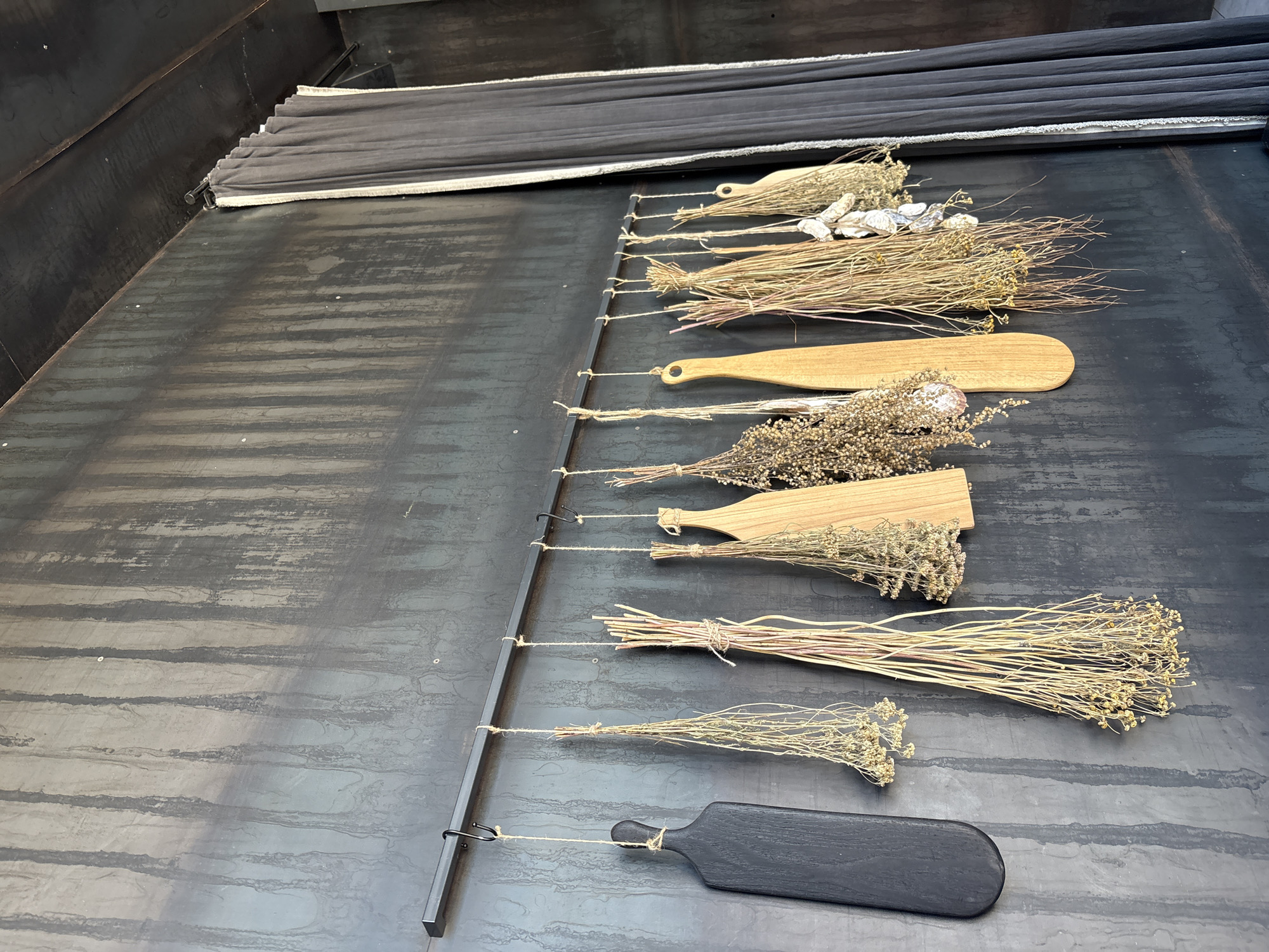 The image displays a wall in Krasnoyarsk, Russia, featuring a distressed dark gray-black surface with vertical light streaks. A horizontal black metal rod is mounted on this wall. From the rod, various kitchen and decorative items are suspended by natural-colored twine and two S-hooks. From left to right, these include a black wooden cutting board, several bunches of dried botanicals (herbs or flowers), three light-colored wooden cutting boards of varying shapes, and a cluster of dried white, papery objects resembling garlic or mushrooms. On the far right, a dark gray curtain with a light-colored trim hangs from a separate rod, partially revealing a matching wall and a dark ceiling beam beyond. No individuals are present in the scene.