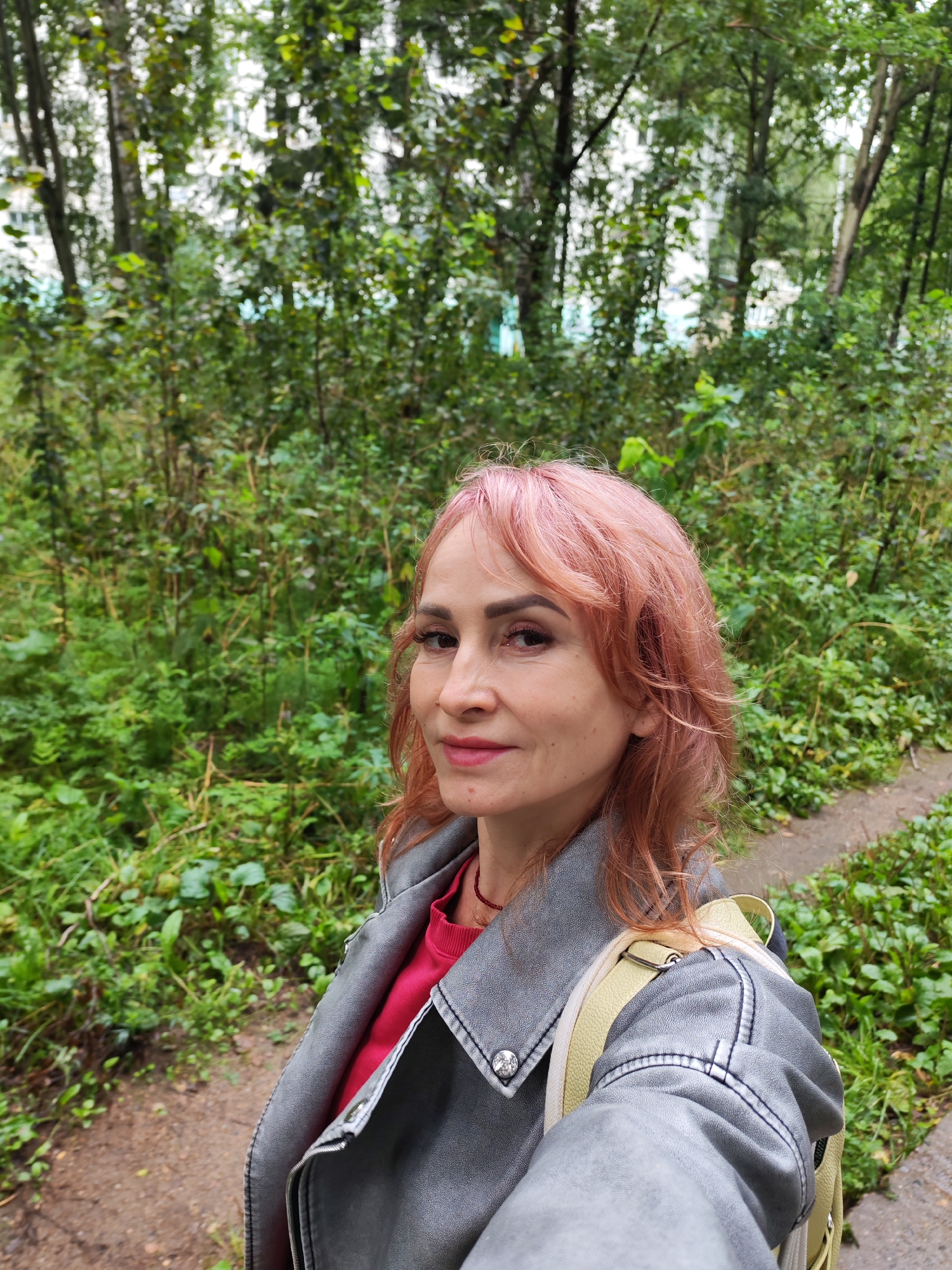 In an outdoor, medium shot, a woman with pink hair stands on a dirt path in a green, wooded area, likely in Chaikovsky, Russia. She is wearing a gray jacket over a pink shirt and a light yellow backpack. She is looking towards the camera with a slight smile. The background consists of dense foliage and trees, suggesting a park or natural setting within the city. The lighting is overcast, indicating a cloudy day.