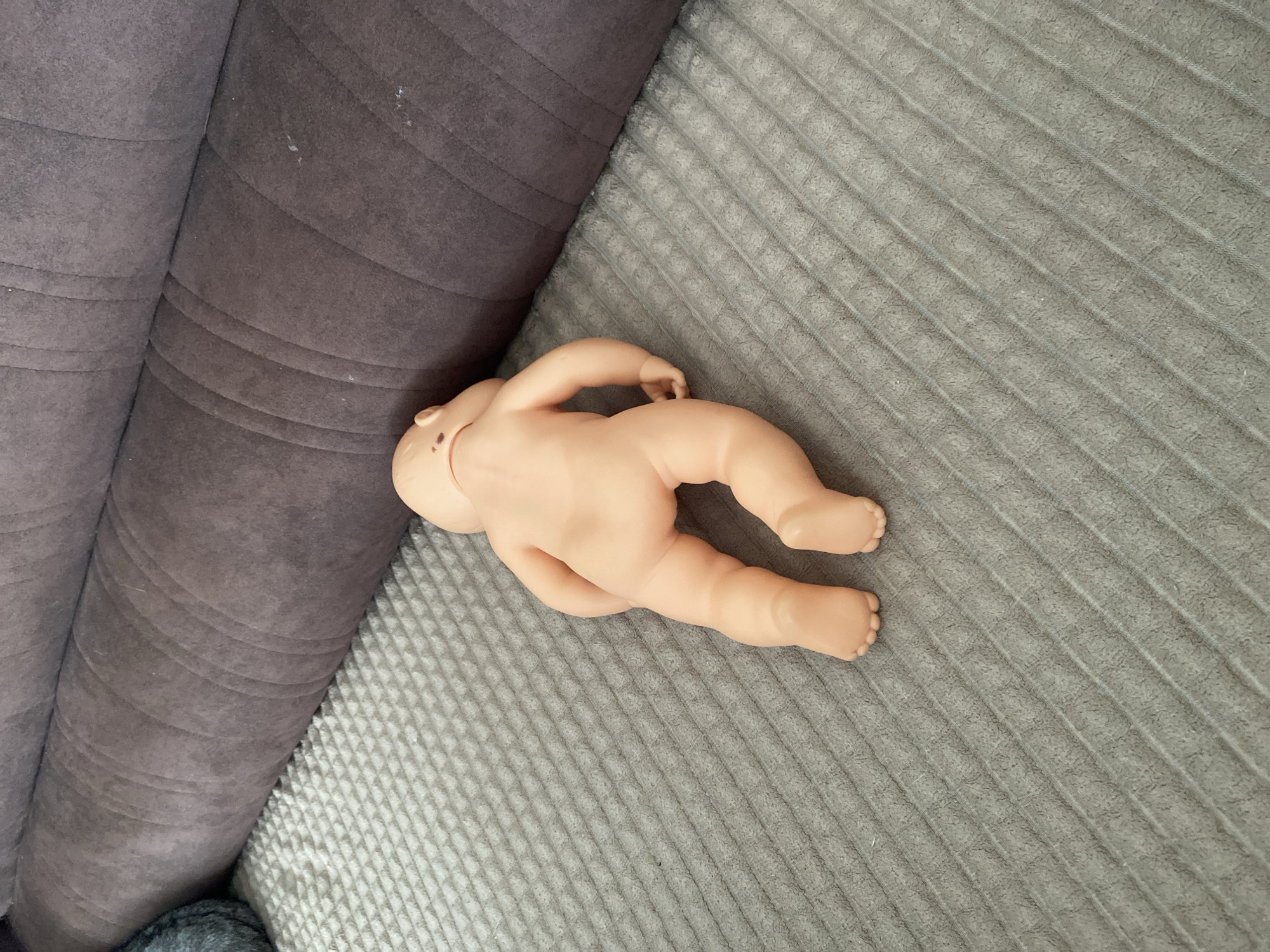 A nude, plastic baby doll is lying face down on a gray, textured blanket. The doll is positioned with its head to the right and its feet pointing towards the left. Behind the doll, a dark purple, plush sofa cushion runs horizontally across the frame. The lighting suggests an indoor setting, with natural light illuminating the scene from an unseen source. There are no other people or objects of significance visible in the image.