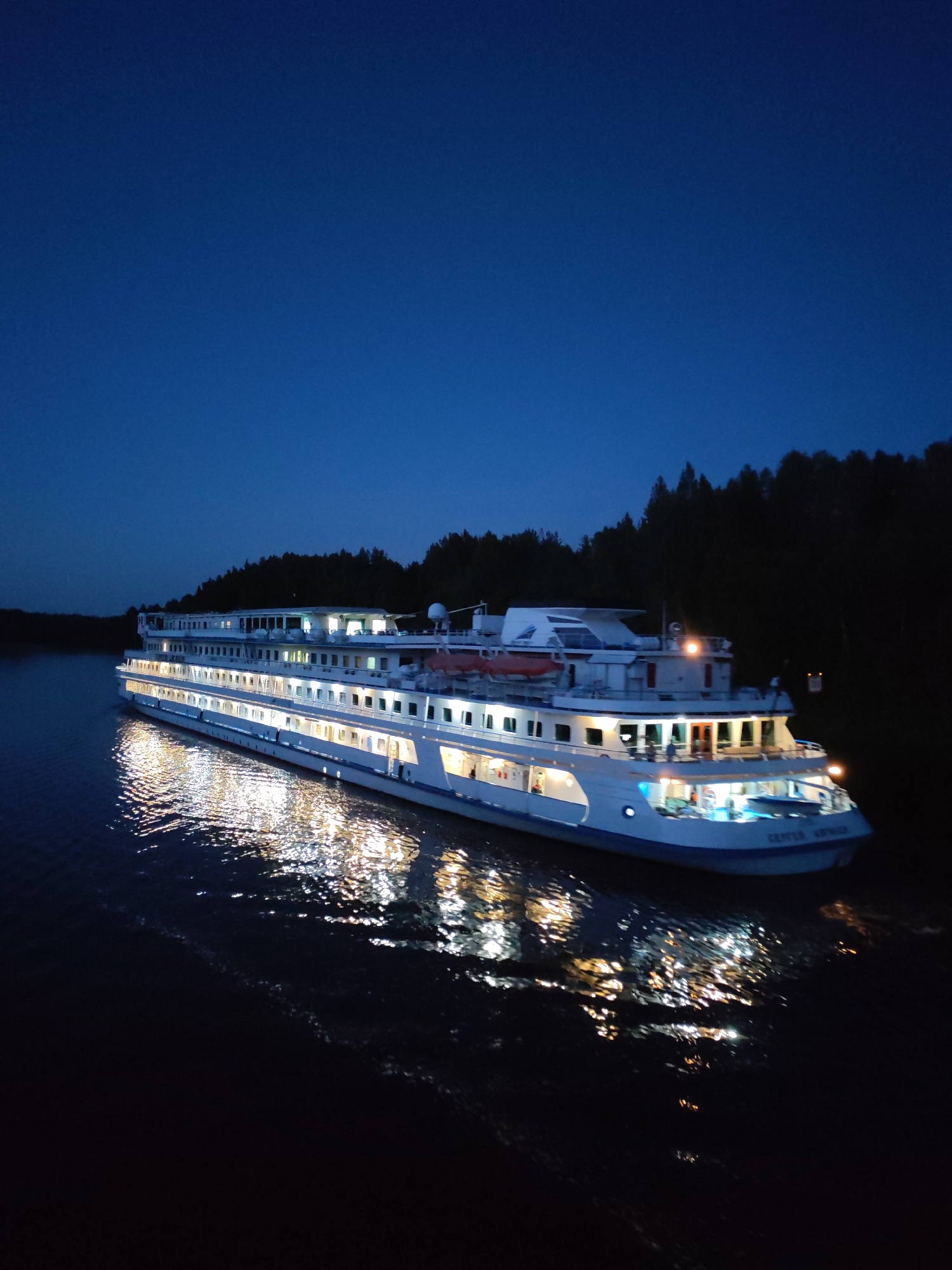 The image depicts a large, illuminated white cruise ship sailing on dark water at dusk or night. The ship has multiple decks, with windows showing warm interior lights. The ship's lights are reflected on the rippling surface of the water, creating a shimmering effect. On the right side of the image, a dense line of dark trees forms the shoreline, with a few lights visible within the treeline. The sky is a deep, clear blue, indicating twilight or night. The name "СЕРГЕЙ АБРАМОВ" is visible on the hull of the ship. The scene appears to be a serene, possibly remote location. While Depo, Russia is mentioned, the specific location within Depo is not identifiable from the visual information. There are no people clearly visible on the ship's exterior decks.