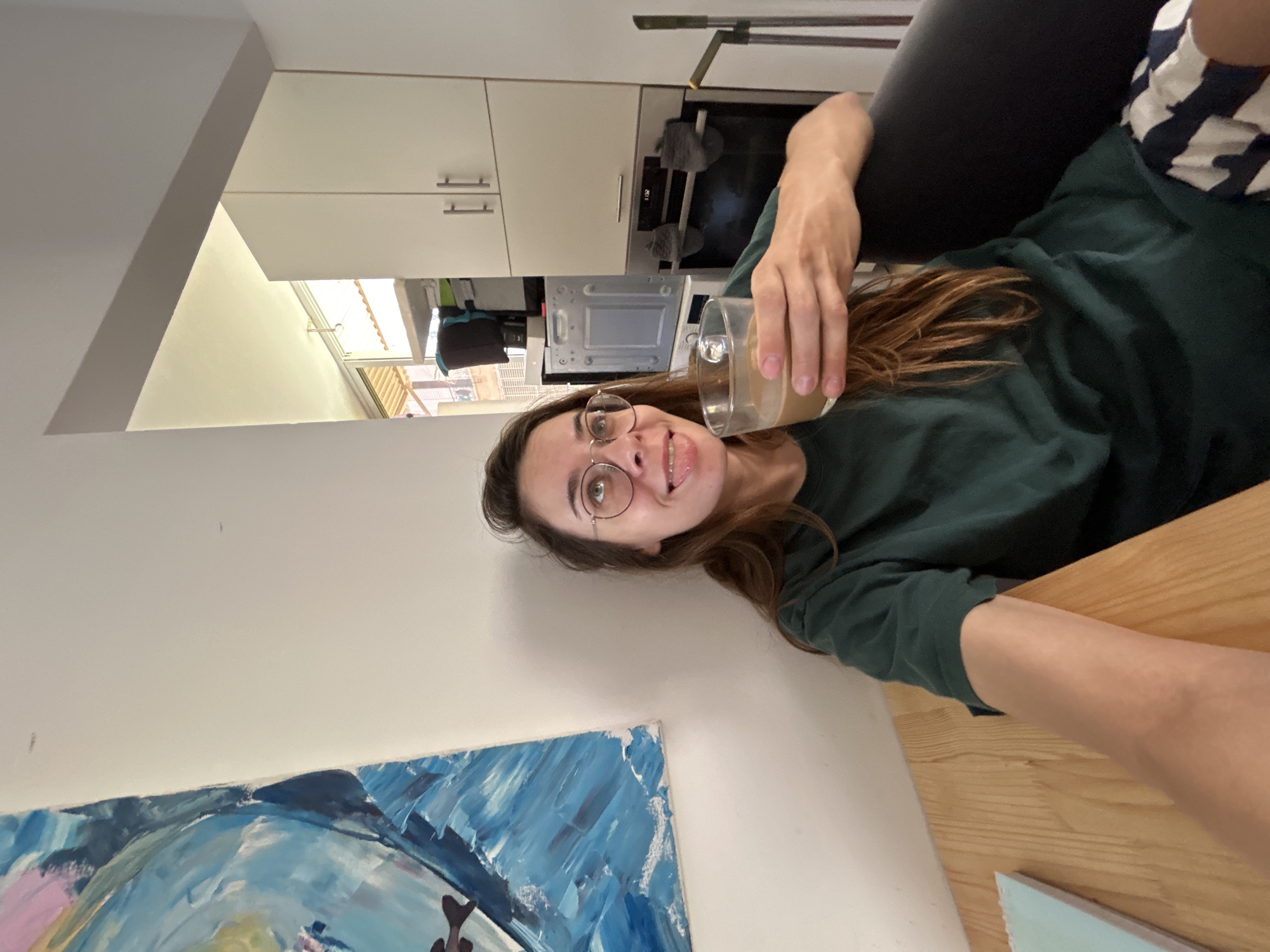 In a residential kitchen in Rishon LeZion, Israel, a young woman with long brown hair and glasses is seated at a wooden table, holding a glass of coffee. She is wearing a dark green t-shirt and striped shorts. To her left is a framed abstract painting with blue and white brushstrokes. The kitchen features white cabinets, a microwave, and an oven with a digital display. The scene is brightly lit, suggesting daylight.