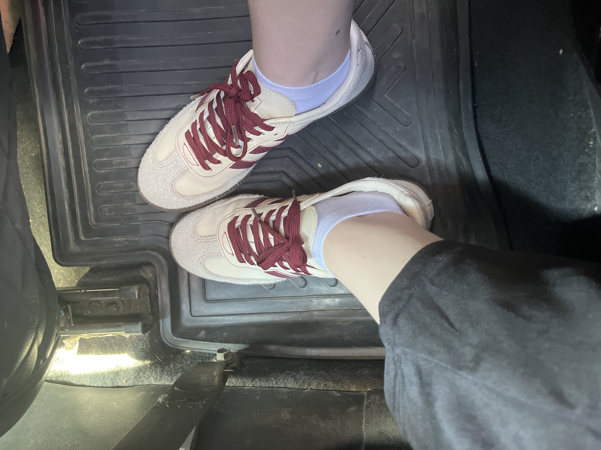 A first-person perspective image shows a person's feet wearing white sneakers with burgundy laces and white ankle socks, standing on a black rubber floor mat inside a vehicle. The left foot is partially obscured by dark gray pants. The vehicle's interior appears to be dimly lit. There is no indication of the location being Sterlitamak, Russia.