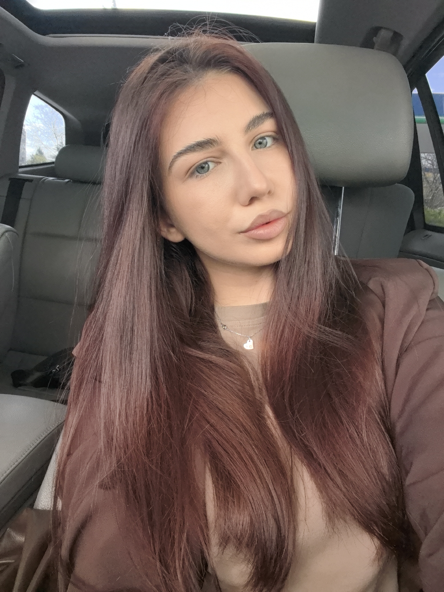 A young woman sits in the driver's seat of a car. She has long, straight auburn hair, fair skin, and blue eyes. She is wearing a brown top and a silver necklace with a heart pendant. The car's interior is grey. The image was likely taken during the day, as indicated by the natural lighting. There is no contextual evidence to suggest the location is Stara Zagora, Bulgaria.