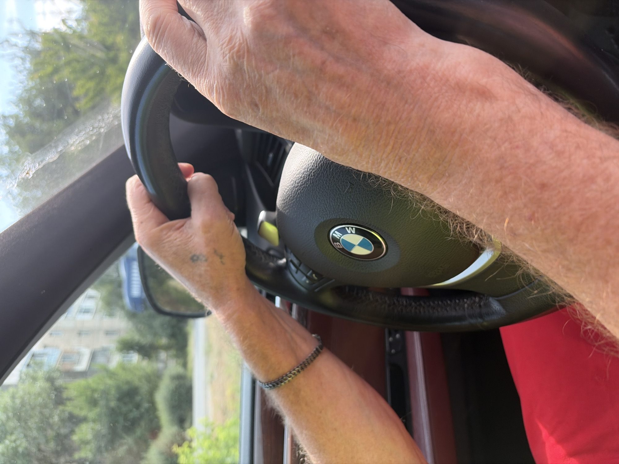 An elderly person's hands are on the steering wheel of a BMW car. The car is in motion, as indicated by the blurred background of trees and buildings. The person is wearing a red shirt and a bracelet on their left wrist. The steering wheel has the BMW logo in the center. The scene appears to be a sunny day.