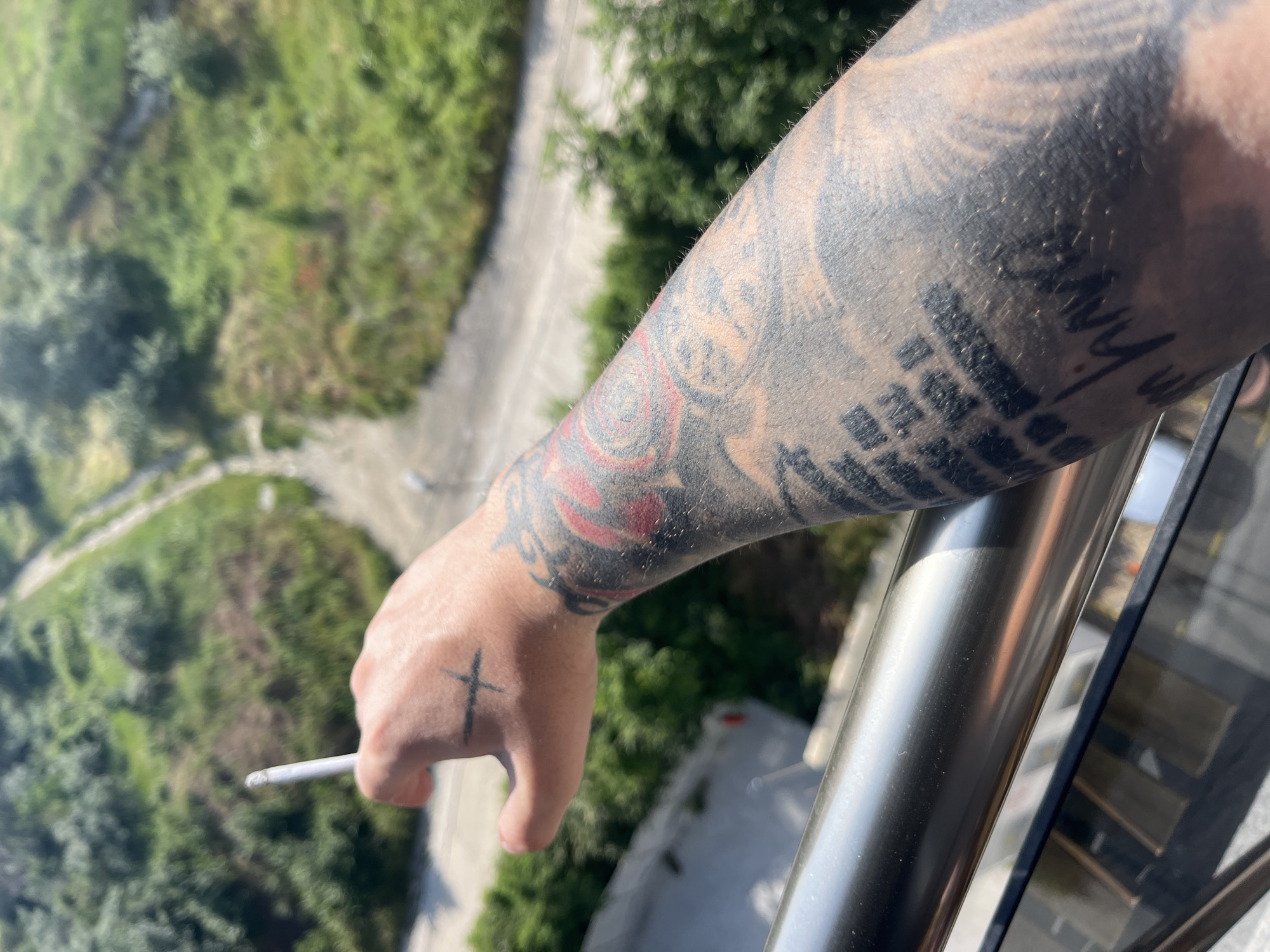 The image shows a close-up of a person's tattooed arm, holding a lit cigarette. The person's hand, which has a black cross tattoo on it, is extended forward. The arm is heavily tattooed with various designs, including what appears to be rose imagery and text. The person is standing on a balcony, indicated by the presence of a metal railing and glass in the foreground. In the background, there is a blurred view of lush greenery and a winding path, suggesting an outdoor setting with a natural landscape. The sunlight indicates it is daytime. The scene appears to be located in the city of Varna, Bulgaria.