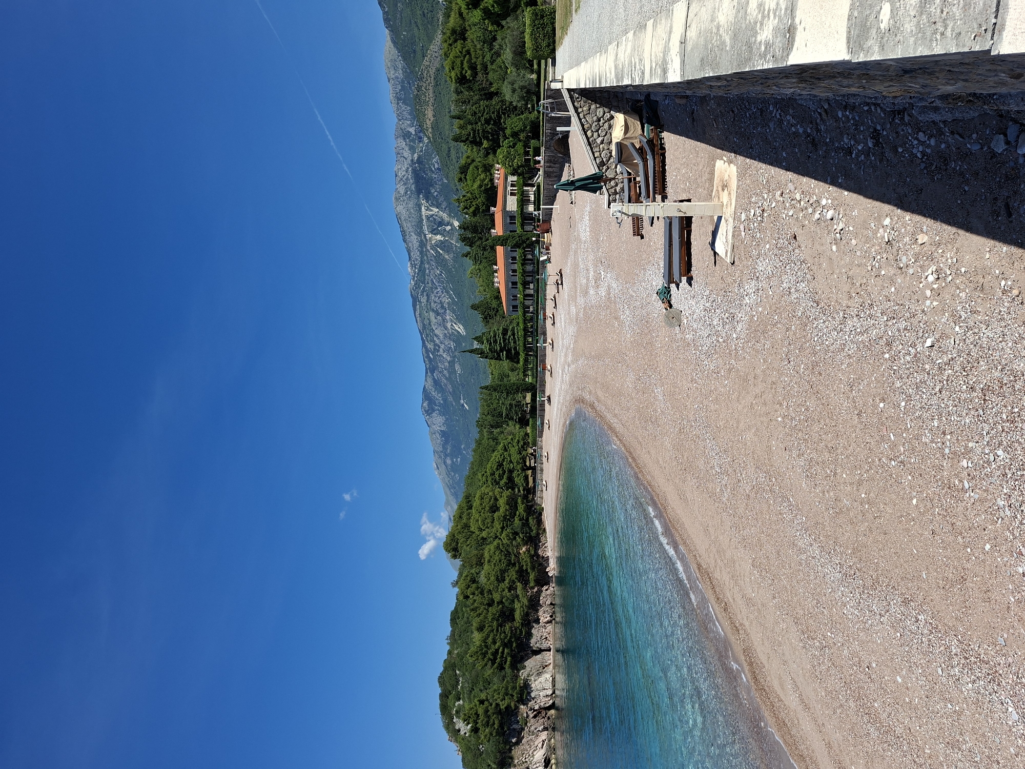 The image shows a view of a pristine beach in Sveti Stefan, Montenegro. The beach is composed of small pebbles and sand, with crystal-clear turquoise water lapping gently at the shore. A curved coastline extends to the left, lined with lush green trees and rocky outcrops. In the background, a large, red-roofed building with a light-colored facade stands amidst dense vegetation. Majestic mountains rise behind the building, their slopes covered in trees and rocky terrain. The sky is a vibrant blue with a few wispy white clouds. Several lounge chairs and umbrellas are scattered on the beach, suggesting a tranquil resort setting. A stone wall runs along the right side of the image, separating the beach from a paved pathway. The overall scene depicts a serene and idyllic coastal landscape.