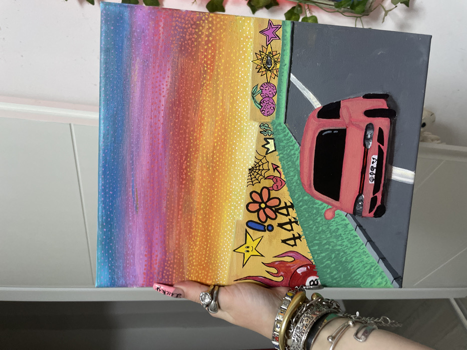 A split-canvas painting shows a juxtaposition of styles. On the left, a vibrant rainbow gradient transitions to yellow with hand-drawn images: stars, cherries, and flowers. The right side features a pink car on a gray road with a white line. The car is framed by green grass. The painting is being held by a hand adorned with multiple bracelets and rings. A cabinet and foliage are visible in the background.