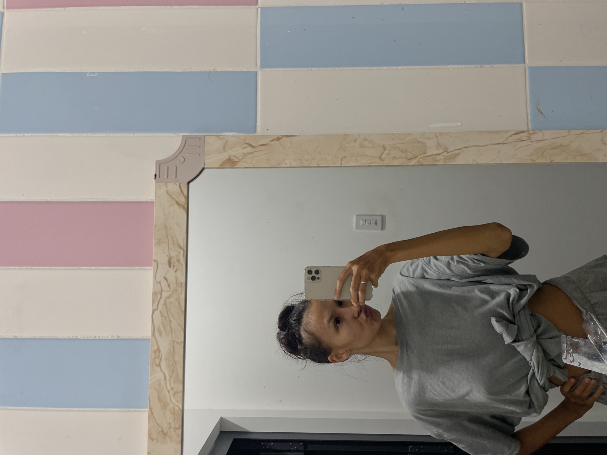 The image shows a person taking a selfie in front of a mirror. The person has their hair pulled back in a bun, and is wearing a grey t-shirt. The phone is held up to their face as if taking a picture in the mirror. The mirror has a decorative frame with a marble-like pattern. Behind the mirror, the wall is tiled with horizontal rows of pastel-colored tiles - pink, white, and blue. In the reflection, part of a window can be seen. The image appears to be taken in a bathroom or similar room. The location, Mũi Né, Vietnam, cannot be definitively determined from the image, but the style of tiling suggests a possible Southeast Asian origin.