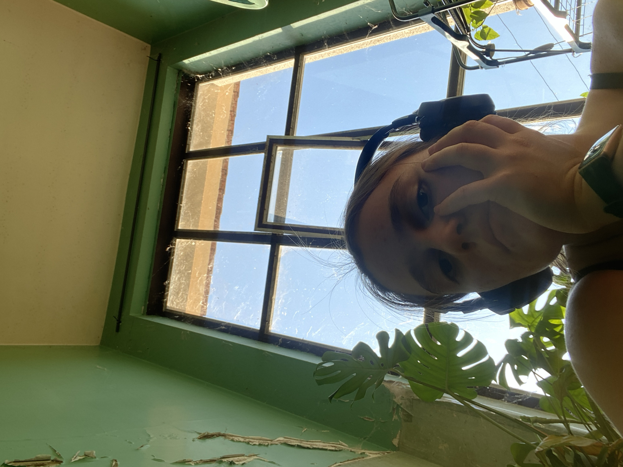 A young woman with light brown hair, wearing black headphones and a black tank top, is looking at the camera while resting her face on her hand. She has a watch on her left wrist. The woman is indoors, in what appears to be a room with peeling green paint on the walls and a window with multiple panes, through which a blue sky is visible. A large-leafed plant is in the foreground, to the left of the woman. A metal shelf unit with plants is visible to the right. The scene is illuminated by natural light coming through the window. The overall impression is that of an urban interior, possibly a room in Belgrade, Serbia, with an aged aesthetic.