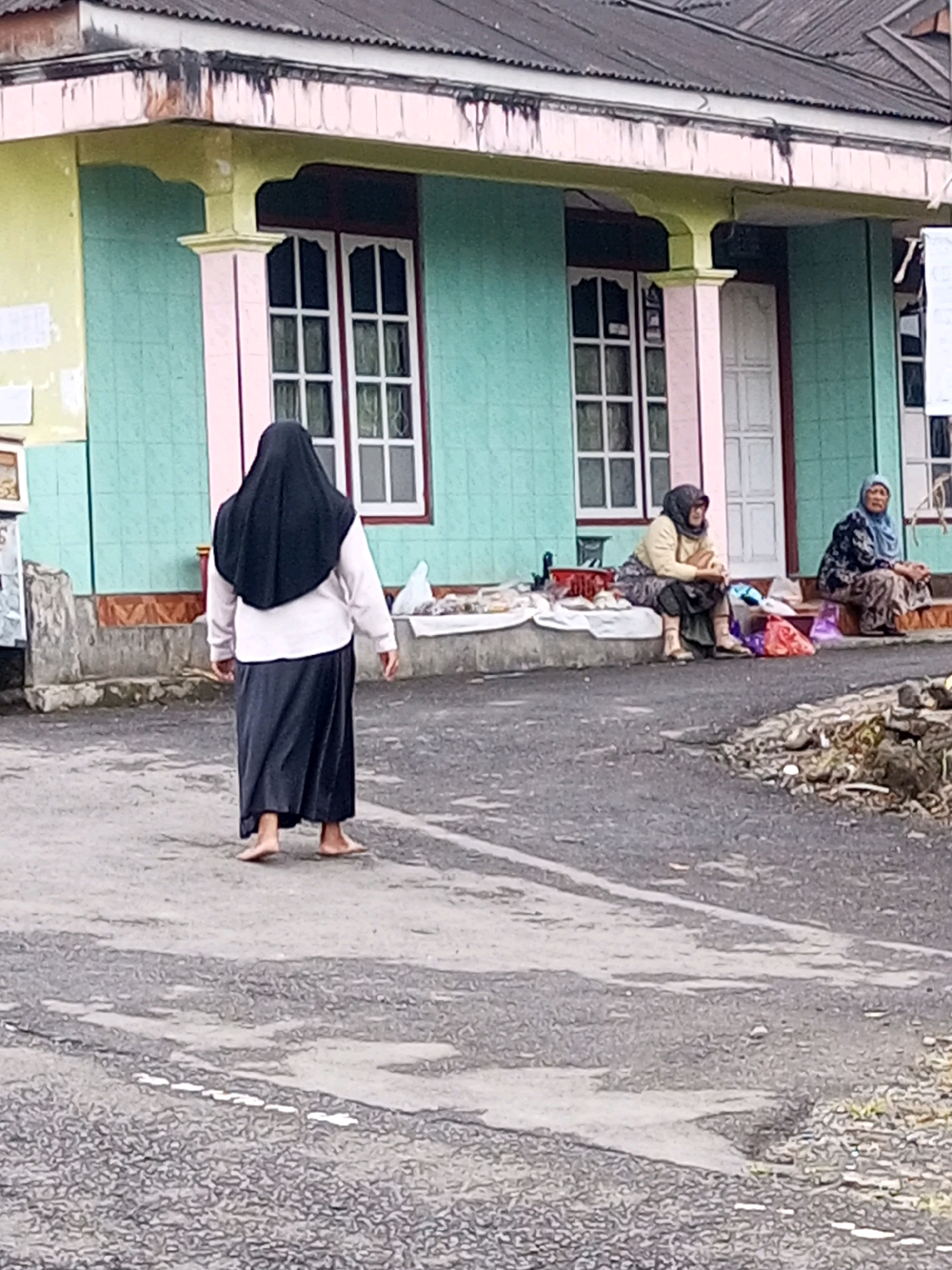 The image depicts a street scene in Keseneng, Indonesia, with a building that has a light green tiled facade, pink trim around the windows and columns, and a light yellow overhang. The roof is corrugated. In front of the building, on the sidewalk, two women are seated. The woman on the left is wearing a yellow jacket and a patterned skirt, with her head covered by a dark scarf. The woman on the right is wearing a blue and black patterned outfit and a light blue headscarf. Both women are facing the building. In the foreground, a third woman is walking away from the camera, barefoot, and is wearing a black hijab and a long dark skirt over a white top. The street is paved with asphalt and shows signs of wear and tear, with some debris on the side of the road. Bags and some items are placed on the sidewalk near the seated women.