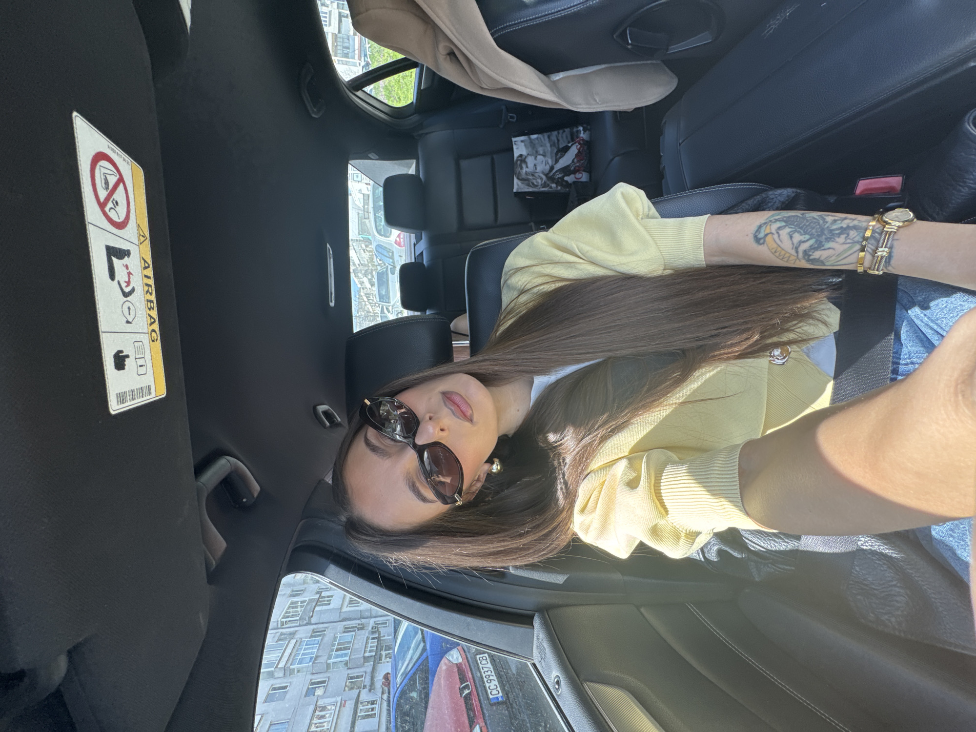 A young woman is in the passenger seat of a car. She has long, brown hair and is wearing sunglasses and a light yellow cardigan. She is also wearing a watch and bracelets on her right arm, which has a tattoo on it. The car is parked on a street in Varna, Bulgaria, and buildings can be seen through the window. A red car, bearing a Bulgarian license plate "CC 9937 CB" is visible in the side mirror. A magazine or a small book is placed on the backseat.
