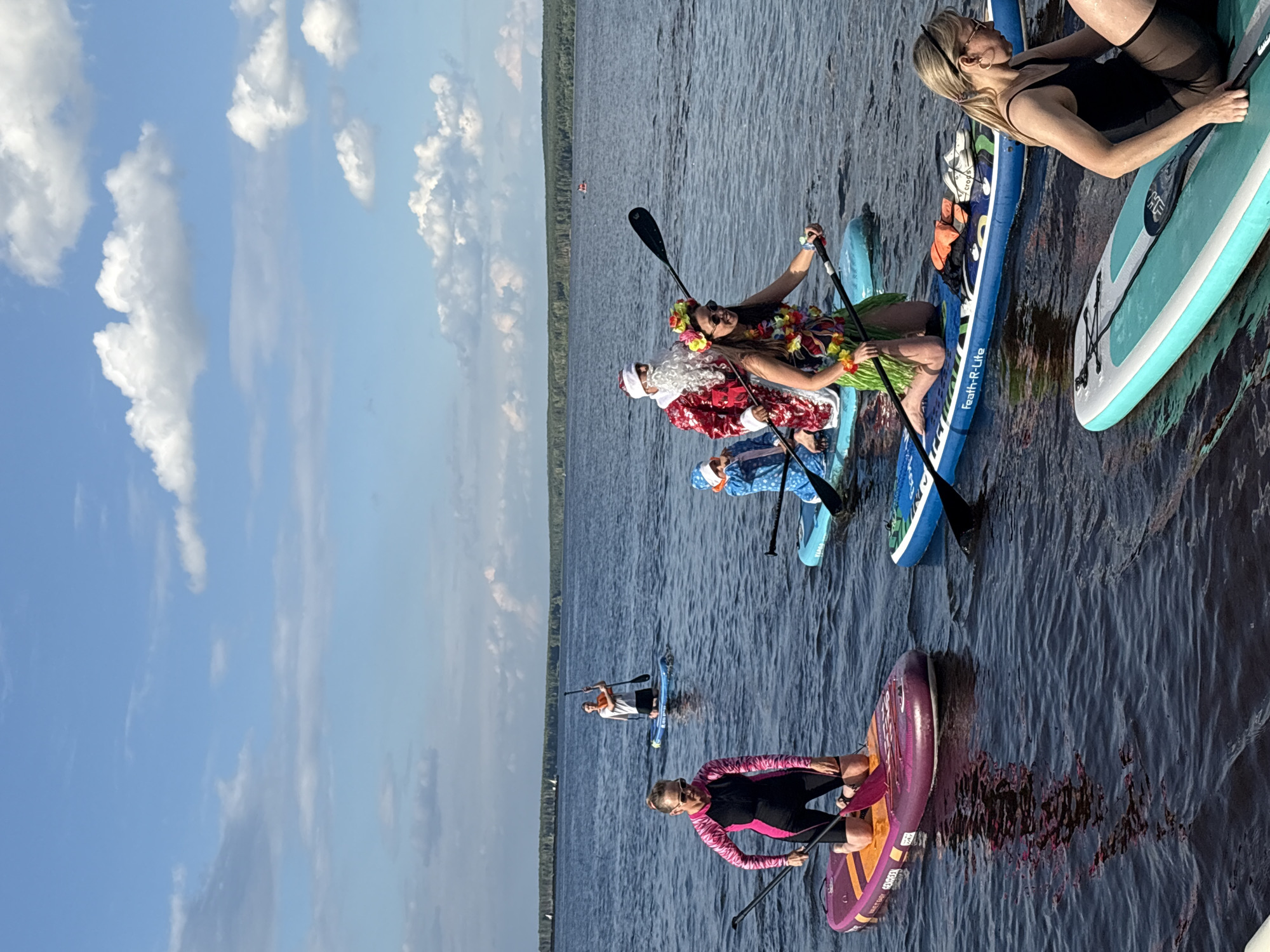 In Chaikovsky, Russia, a group of people are paddleboarding on a large body of water, likely the Kama River, under a partly cloudy sky. The scene takes place during daylight, with the sun illuminating the water and the surrounding tree-lined shore. Several individuals are on paddleboards, some kneeling and others in a standing or seated position, holding paddles. One person is dressed as Santa Claus, complete with a red suit and white beard, on a paddleboard with a child in a blue snowsuit. Another person on a paddleboard is wearing a Hawaiian-themed outfit with a lei and a grass skirt, and a third person is dressed in a black swimsuit and sunglasses. The water is dark blue and shows reflections of the sky and the paddleboards. In the distance, a few sailboats are visible on the horizon, and the shoreline is densely covered with trees.