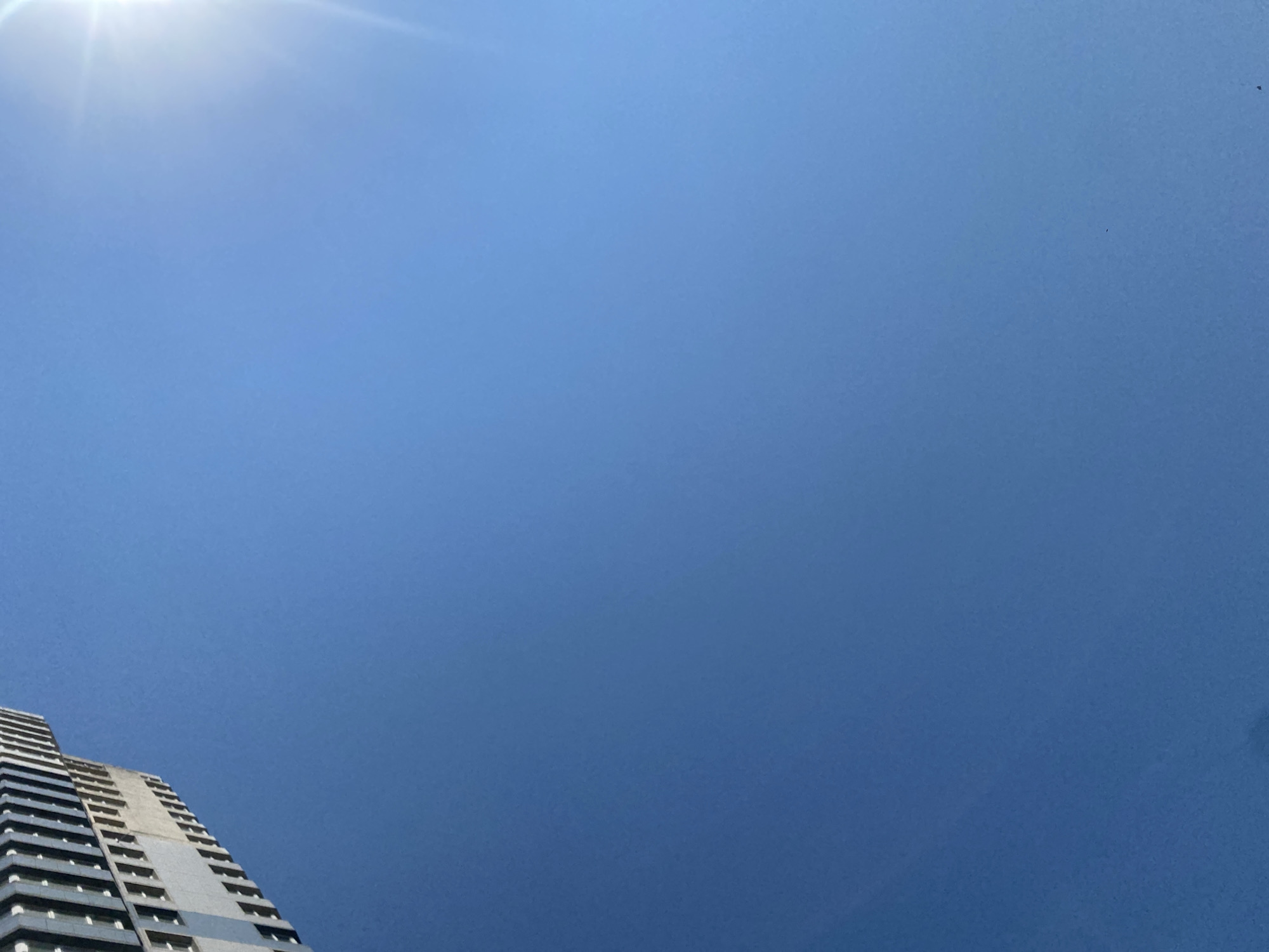 The image is a low-angle shot looking up at a clear blue sky with a bright sun visible on the right side. In the upper left quadrant, a portion of a tall, modern building with a facade of windows is visible, angled against the sky. The building appears to be constructed with concrete and glass. The sky is unobstructed and appears to be a bright, sunny day. There are no people or animals visible in the image. The scene is likely an urban environment, given the presence of the building. The location is identified as Chornomorsk, Ukraine.