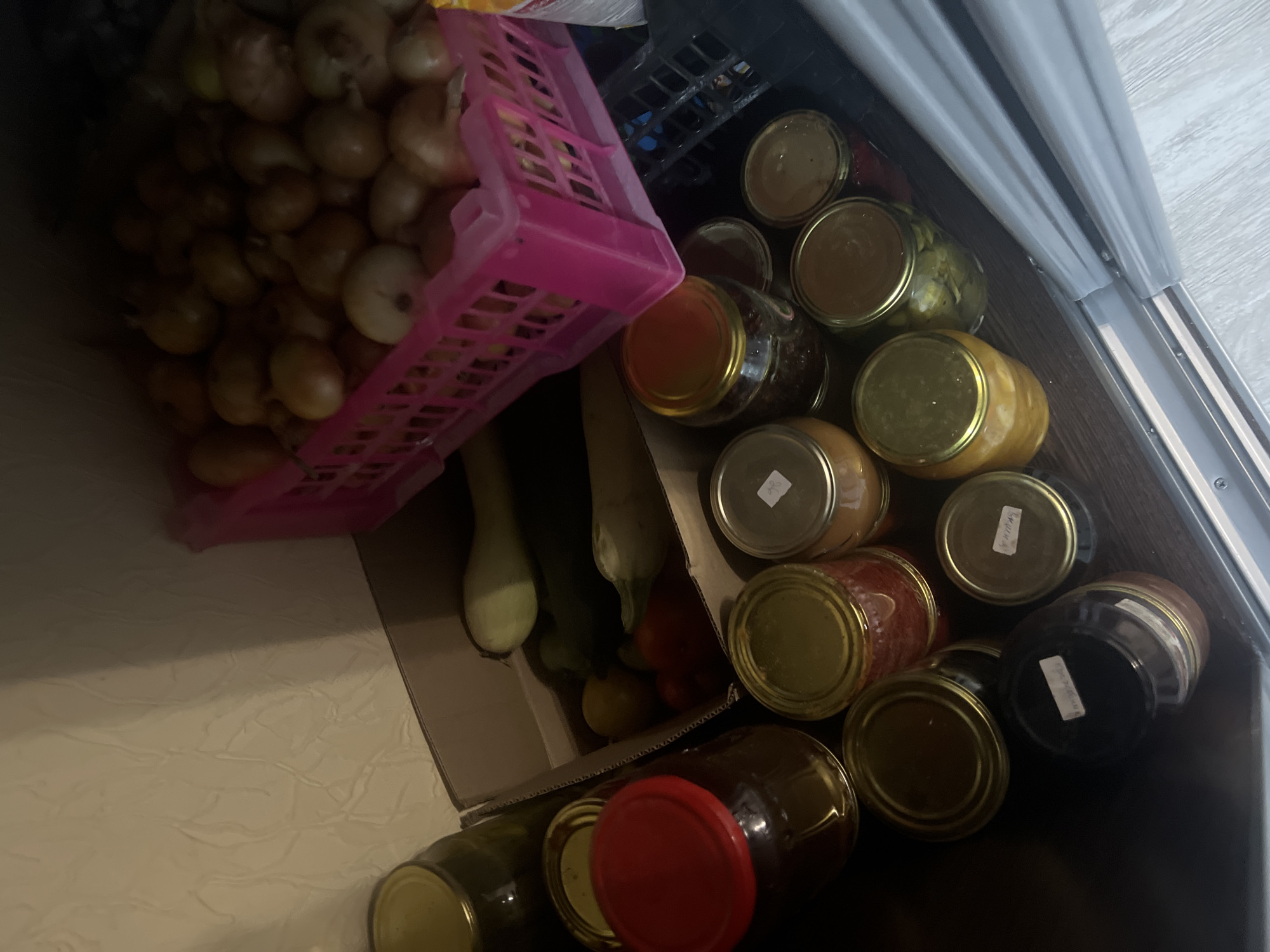 A collection of preserved food items and produce is displayed on a dark wooden surface in what appears to be a pantry or storage area. In the background, a pink plastic crate overflows with a large quantity of onions. To the left, a cardboard box contains several green zucchini and red bell peppers. A large glass jar with a red lid is prominent in the foreground, alongside several smaller glass jars with gold-colored lids, some containing visible food items such as pickles and preserves. The environment suggests a domestic setting where food is being stored, likely for later consumption. The image does not contain any information that would allow for its localization to the city of Surgut, Russia.